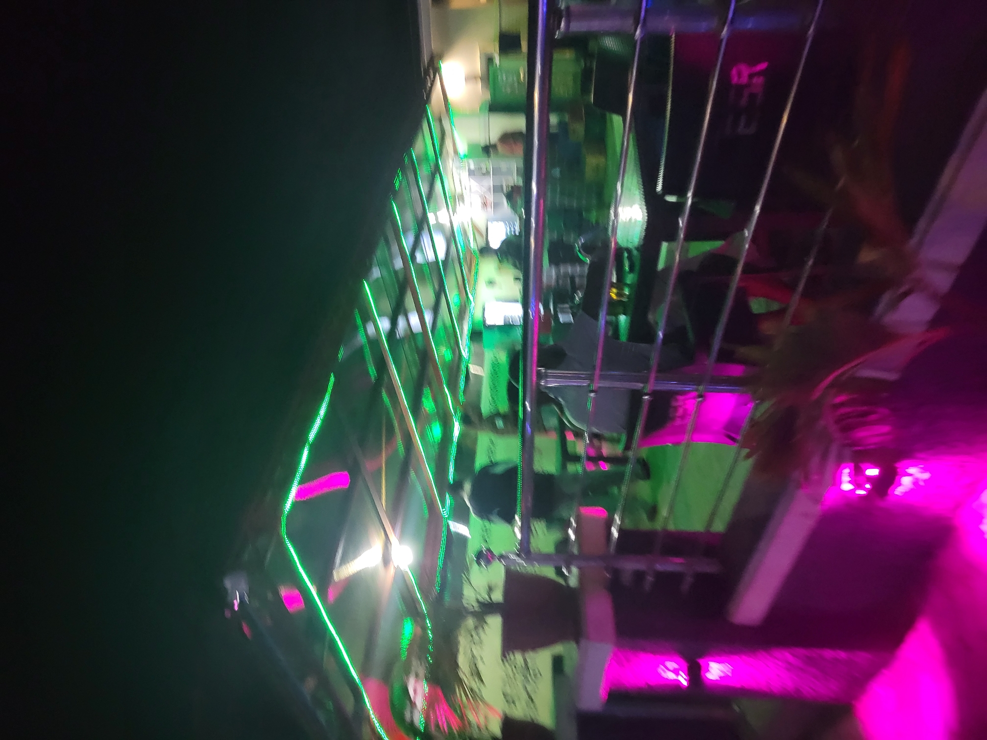 This image depicts a vibrant, dimly lit indoor setting, likely a nightclub, bar, or event space, in Ibo, Nigeria. The scene is dominated by striking green and magenta/purple LED strip lights that crisscross the frame, creating luminous streaks due to motion blur or a long exposure. These lights reflect off vertical, metallic-looking bars, suggesting a decorative enclosure or stage setup.
In the background, behind the reflective bars, several individuals are visible. One person appears to be operating a DJ console or control panel, indicating that music is being played or performed. Other figures are less distinct, possibly patrons enjoying the atmosphere. The overall activity seems to be a lively social gathering or party.
The time of day is clearly nighttime, given the complete reliance on artificial lighting. The combination of intense colored lights and reflective surfaces creates an energetic and festive mood. Some blurred foliage, possibly indoor plants, can be discerned, adding to the decor. Faint, blurry white text, which might read "ESR" or similar, is barely visible on a dark surface in the background, but it is largely illegible due to the image's overall blur and low light conditions. The image conveys a dynamic and celebratory ambiance.