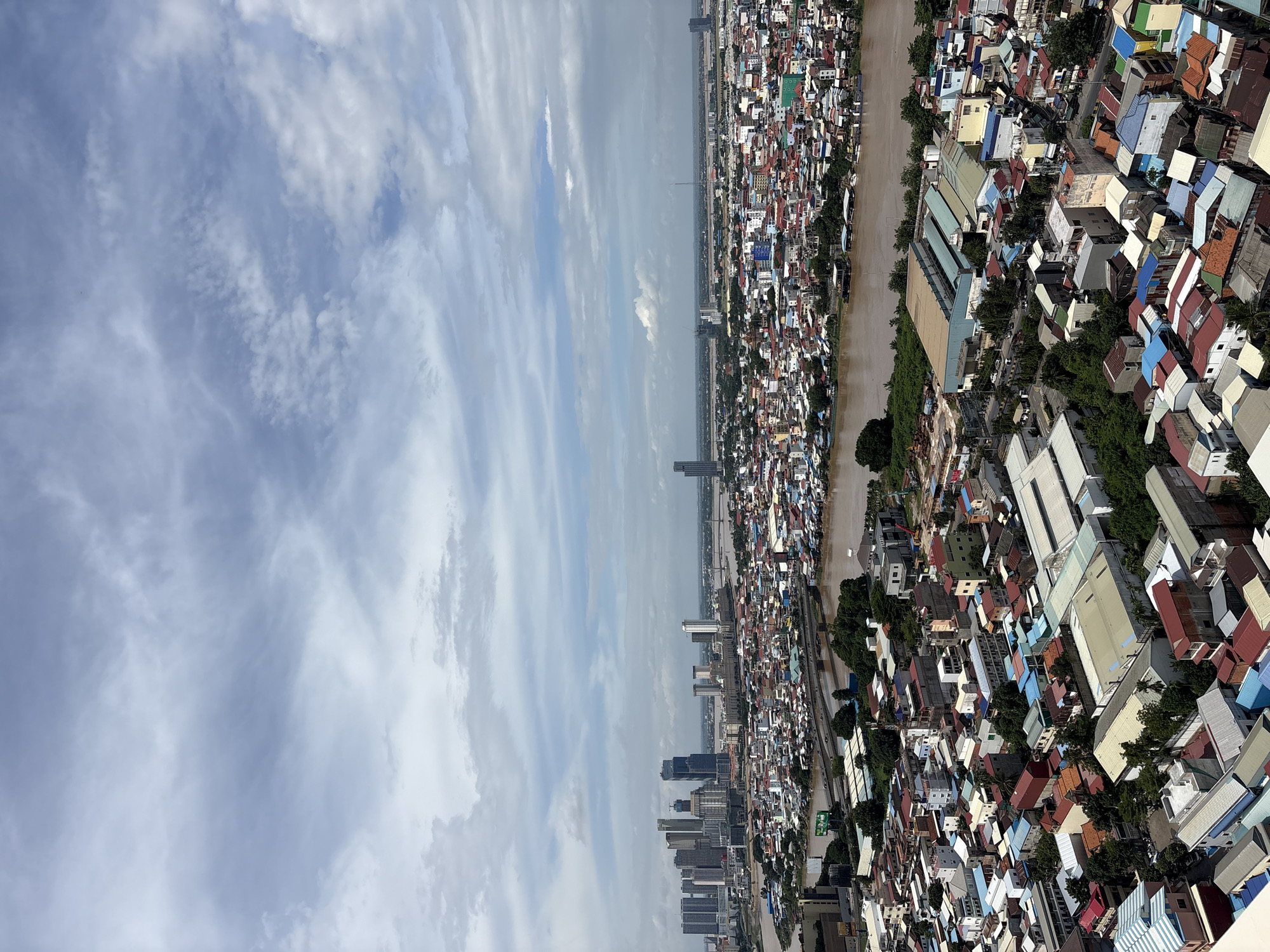 This is a high-angle aerial view of a densely populated urban area on the banks of a wide, muddy river, likely within the Khan Mean Chey district of Phnom Penh, Cambodia. The cityscape is characterized by a mix of low-rise residential buildings with colorful, corrugated metal roofs and some multi-story modern buildings, including a cluster of skyscrapers in the distance. The river, which appears to be the Mekong River or one of its tributaries, dominates the middle ground, with a network of bridges spanning it, connecting different parts of the city. Boats are visible on the river, suggesting continued use for transportation and possibly habitation. The foreground is filled with numerous houses and smaller structures, many with brightly colored roofs, tightly packed together. Vegetation, primarily trees, is interspersed throughout the residential areas and along the riverbanks. The sky is partly cloudy, with soft white clouds against a pale blue background, indicating a day with diffused sunlight. No individuals are clearly discernible in the image, and the focus is on the urban landscape and its infrastructure.