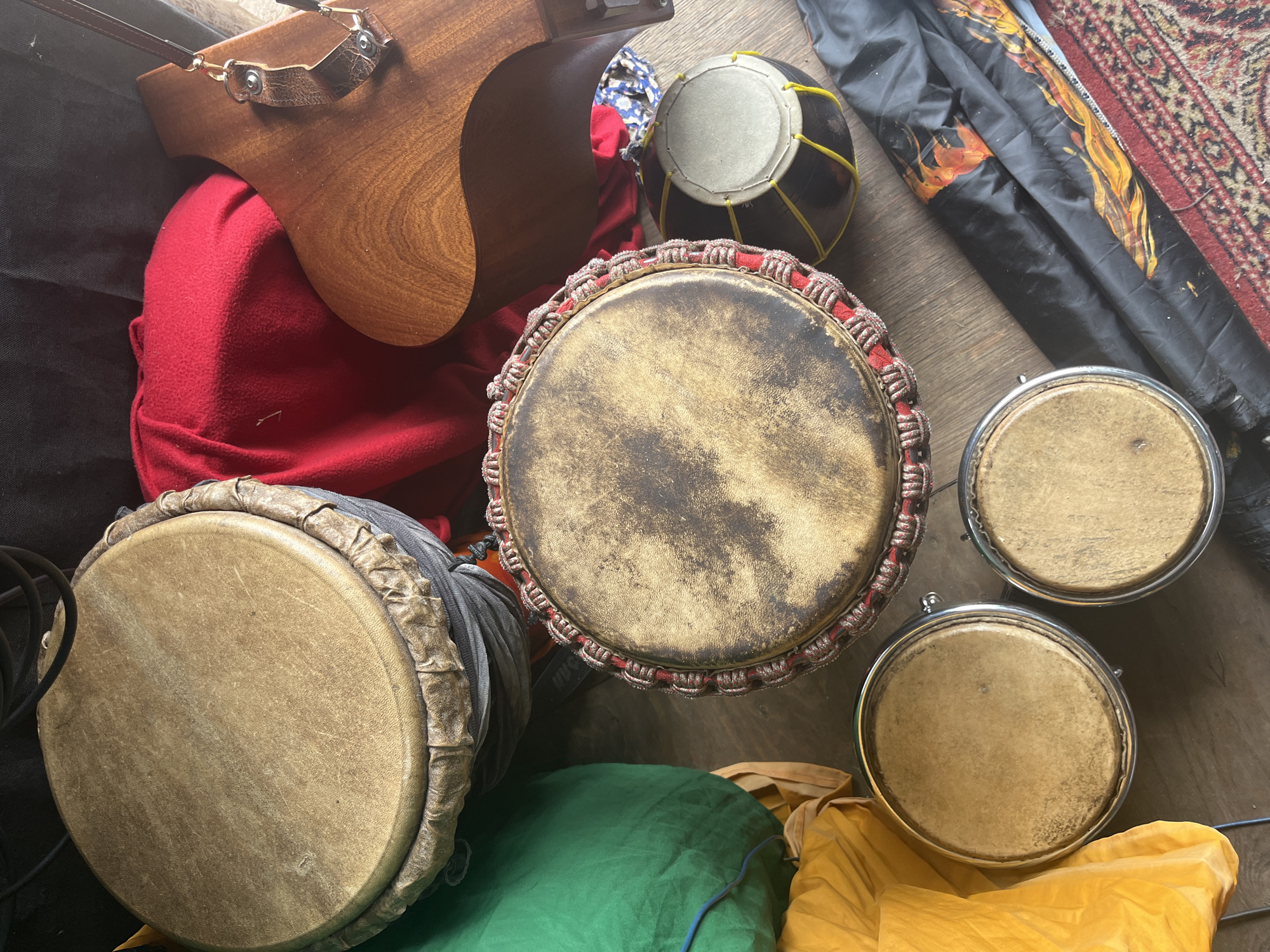 An overhead, high-angle shot captures a collection of drums and other items on a wooden floor. In the upper left, a large djembe drum with a weathered, tan hide head is positioned vertically. To its right, a red fabric is draped over a dark object. A portion of a wooden instrument, possibly a guitar body, is visible in the upper right corner, with a leather strap attached.
Below the large djembe, a green fabric bundle rests on the floor. To the right of the green bundle, a medium-sized drum with a worn, light brown hide head and a decorative red and white rope trim is prominently displayed. Next to it, a smaller black drum with a light-colored head is secured with yellow cord.
In the lower portion of the image, two smaller drums, possibly bongos or congas, with tan hide heads and silver hardware, are positioned side-by-side. To the left of these drums, a bright yellow fabric is folded. A black fabric with a flame design and a red patterned rug are visible in the lower right. The lighting is natural and the scene appears to be indoors.