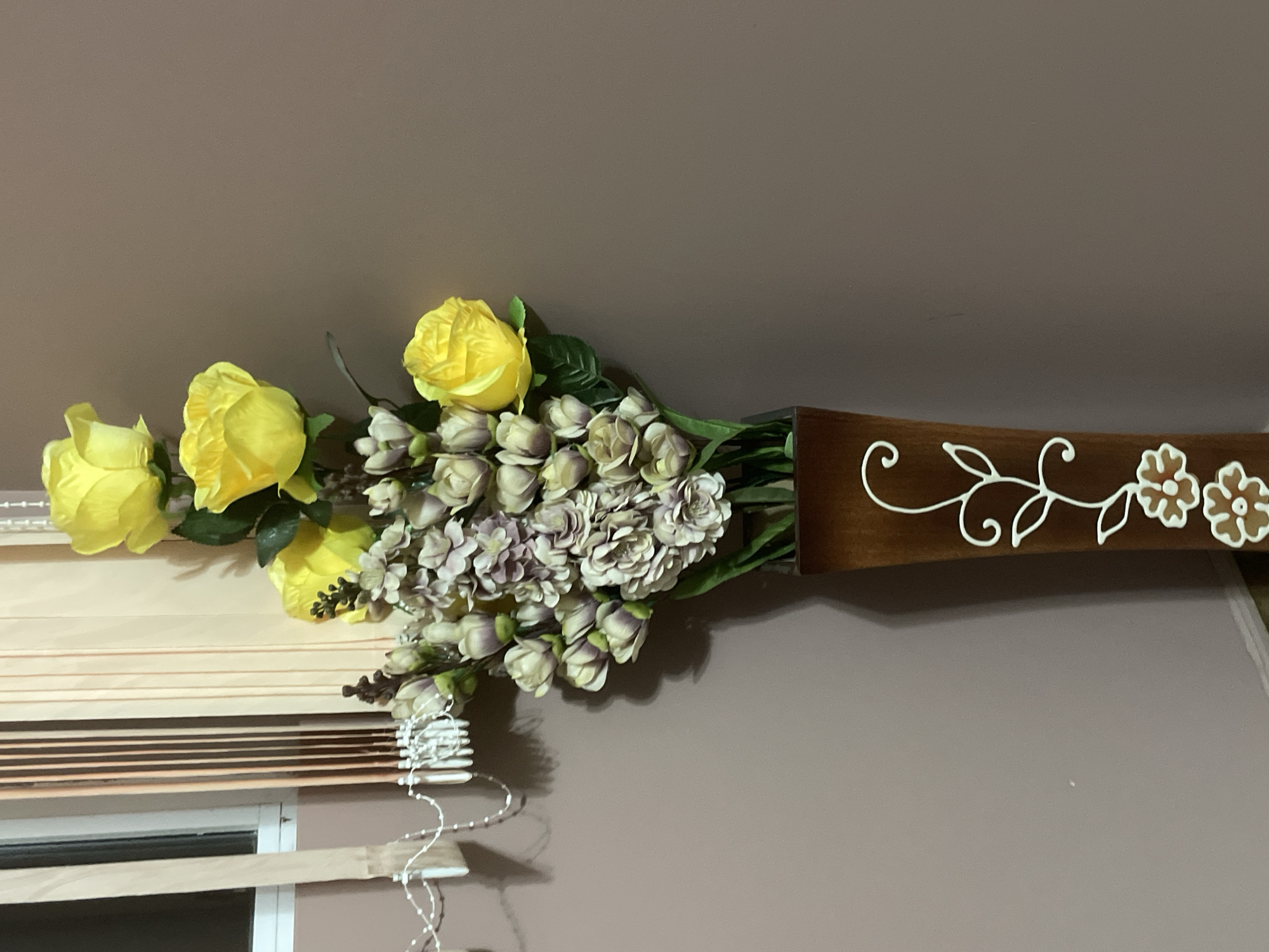 The image captures an indoor scene featuring a decorative wall-mounted floral arrangement. The central subject is a bouquet of artificial flowers held within a dark brown, possibly wooden, rectangular vase or holder. The bouquet primarily consists of several vibrant yellow roses, complemented by numerous smaller, muted purple, grey, or beige flowers, some appearing as closed buds.
The vase is embellished with white decorative scrollwork and two stylized white and brown floral motifs on its right side. The background behind the arrangement is a plain, solid light brown or taupe-colored wall.
In the bottom left portion of the image, part of a window is visible, covered by light-colored horizontal blinds, likely beige, cream, or light wood. The operating strings for the blinds are also discernible.
No people are present in the image, and no specific activity is taking place; the scene depicts a static decorative display. The time of day or weather conditions cannot be determined from this indoor shot. There is no visible text anywhere in the image. The location is contextualized as Ihtiman, Bulgaria, though no specific visual cues in the image indicate this.