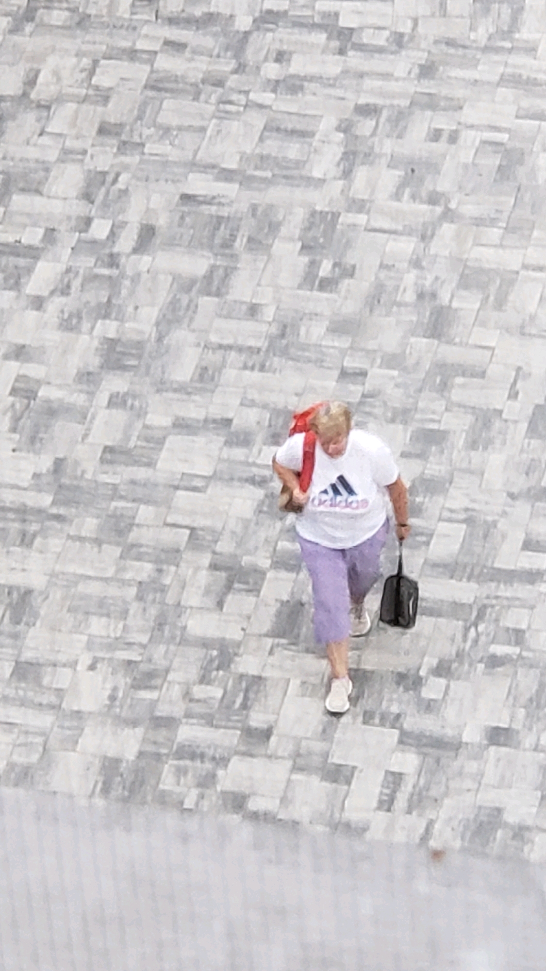 An adult woman with blonde hair, wearing a white Adidas t-shirt and light purple capri pants, walks across a paved area. She carries a red backpack over her left shoulder and a black bag in her right hand. The paving stones are rectangular and arranged in a diagonal, interlocking pattern with varying shades of gray and white. The scene is viewed from above, suggesting an elevated vantage point. There is no identifiable signage or architectural detail to confirm the location as Prešov, Slovakia.