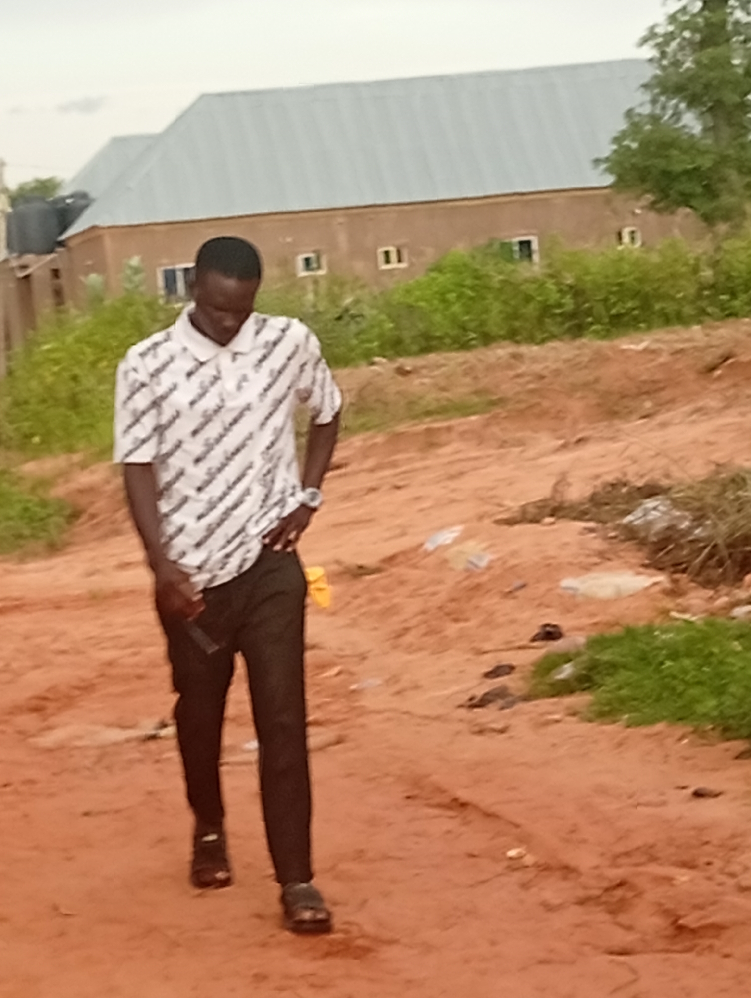 A full-body shot, captured outdoors, depicts a young Black male walking on a dirt path in Tumfure, Nigeria. He is wearing a short-sleeved, white polo shirt with a black all-over pattern and black trousers. His left hand is on his hip, and his right arm is slightly bent at the elbow, with his hand near his pocket. He is wearing dark-colored sandals. In the background, a long, single-story building with a corrugated metal roof is visible, surrounded by green foliage. The ground is composed of reddish-brown dirt with some scattered debris. The sky is overcast.