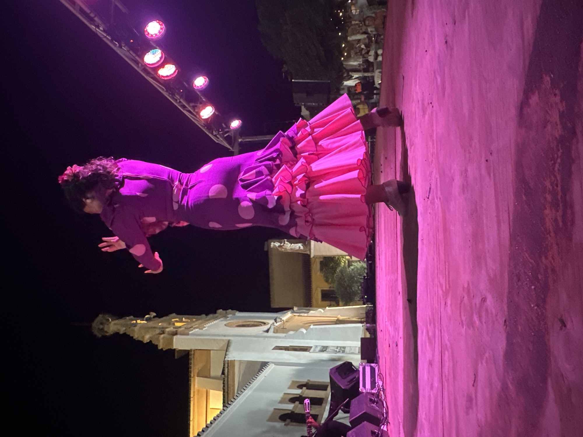 A flamenco dancer performs on a stage at night. The dancer is wearing a vibrant purple dress with large white polka dots and a pink ruffled hem. Her arms are raised and her hands are positioned in a graceful pose. The stage is illuminated by bright pink stage lighting, which also casts a shadow of the dancer on the floor. In the background, a white building with arched windows, possibly a church, is visible. A string of colorful lights hangs above and to the right of the stage, and a few people are seated in the background, observing the performance. The overall scene suggests an outdoor cultural event taking place in San José Del Valle, Spain.