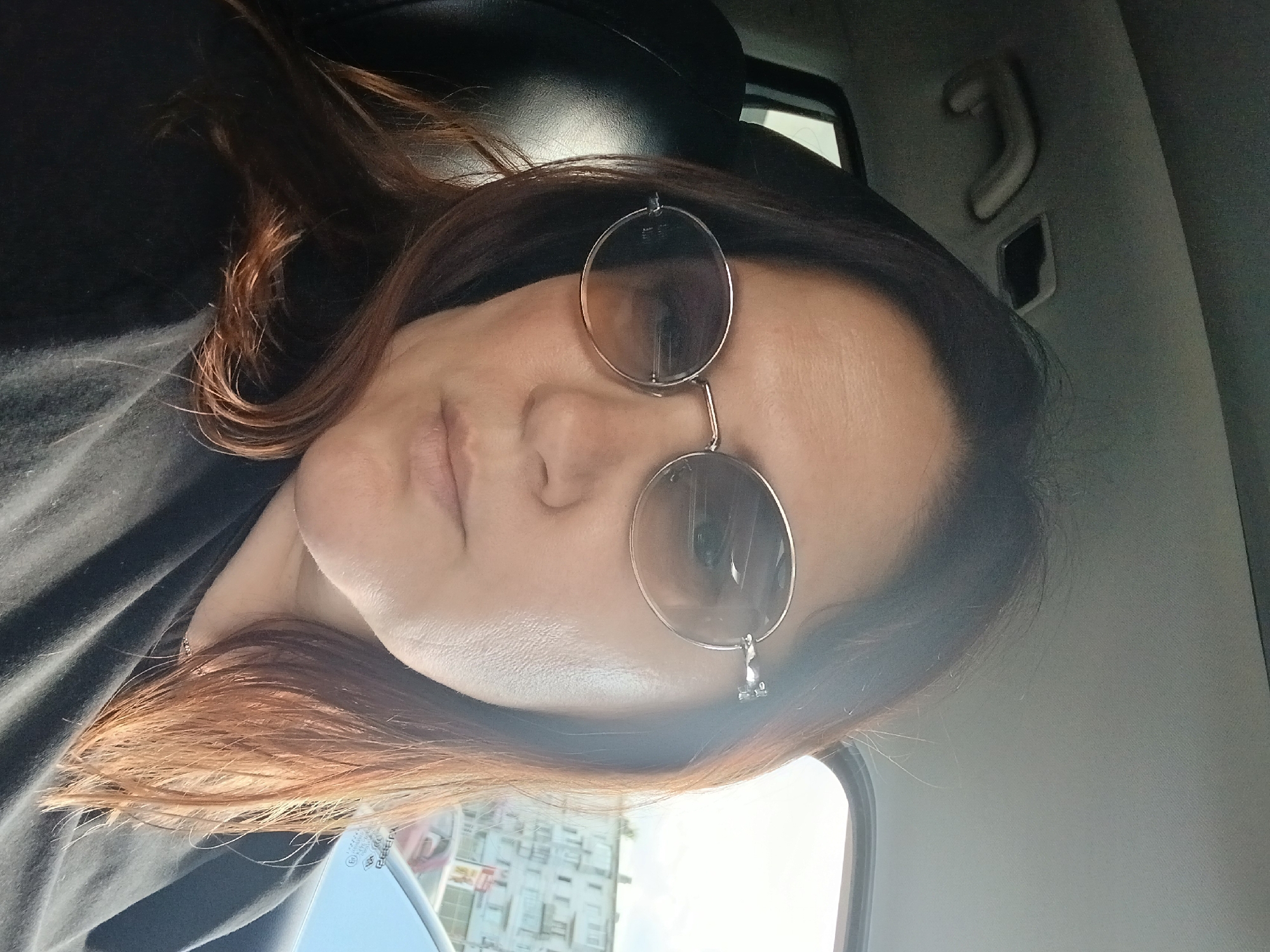 A close-up, sideways shot captures a woman wearing round, silver-rimmed glasses. She is indoors, likely in a car, as indicated by the car's interior visible in the background, including a handle and a portion of the ceiling. Her dark hair, with reddish highlights, is visible around her face and shoulders, obscuring part of the frame. The lighting suggests it might be daytime, with natural light filtering in from outside. The woman's expression is neutral, and her eyes are hidden behind the tinted lenses of her glasses. The overall mood is casual and personal.