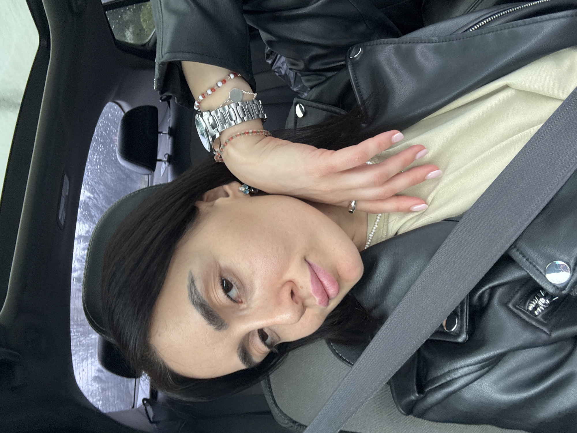 A female individual is depicted taking a selfie inside a dark-colored vehicle in Tbilisi, Georgia, during wet weather conditions. The individual, with dark hair, dark eyebrows, and light-colored skin, is seen wearing a black leather jacket over a light beige top, secured by a grey seatbelt across her chest. Her lips are a light pink. On her left wrist, she wears a silver-toned watch, multiple beaded bracelets (red, white, and orange tones), and a delicate chain bracelet with a small silver circular charm. A silver-toned ring is visible on her right ring finger. A small, multi-colored earring (blue/green tones) is present in her left ear, and a pearl necklace is partially visible beneath her top. Her right hand is positioned with fingers slightly curled, resting on her left forearm. Her fingernails are painted a light pink shade. The car interior features dark upholstery and a visible sunroof. Through the rear and side windows, rain droplets are clearly visible, indicating precipitation, with blurred outdoor foliage in the background.