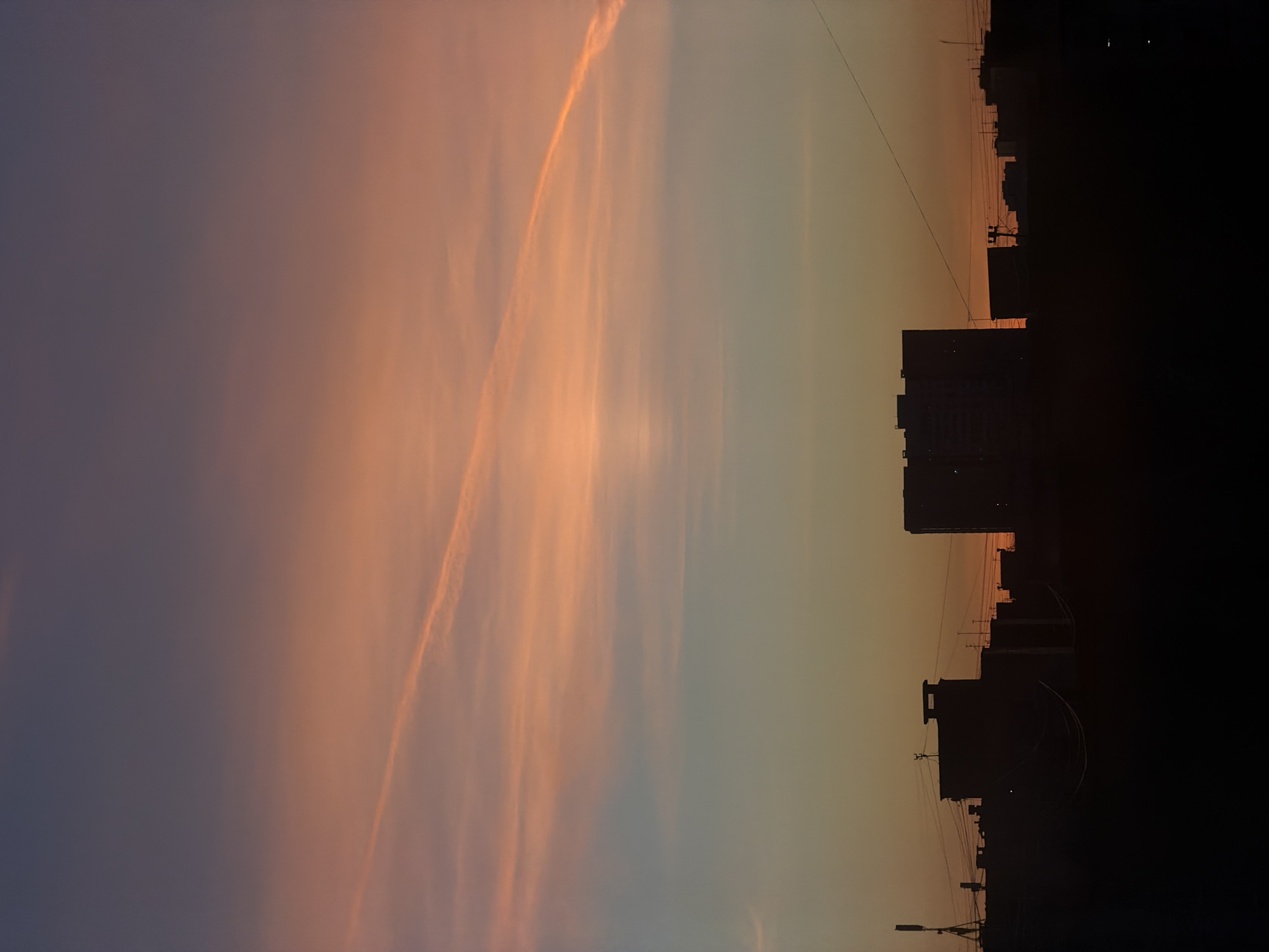 The image displays an urban twilight scene in the city of Shul'gino, Russia, observed during either sunrise or sunset. The sky occupies approximately three-quarters of the frame, exhibiting a distinct color gradient: dark purplish-blue at the zenith, transitioning to a prominent orange-pink hue across the mid-sky, and fading to a lighter orange-yellow near the horizon. Thin, wispy cloud formations are visible within the orange-pink strata. A single, linear condensation trail extends diagonally from the upper-left to the mid-right, appearing brighter against the surrounding sky.
The lower quarter of the image features the silhouette of an urban skyline. Multiple multi-story buildings are present as dark, indistinct forms. A tall, rectangular building is visible on the far left, while a large, blocky structure dominates the central skyline. Several smaller, less defined buildings are scattered across the horizon line to the right. Thin, dark lines, consistent with power or communication cables, traverse horizontally across the mid-ground, connecting between unseen points and visible building structures. A tripod-like antenna or mast is faintly discernible on a rooftop towards the left side of the frame. No individuals, specific actions, or direct interactions are identifiable within the scene.