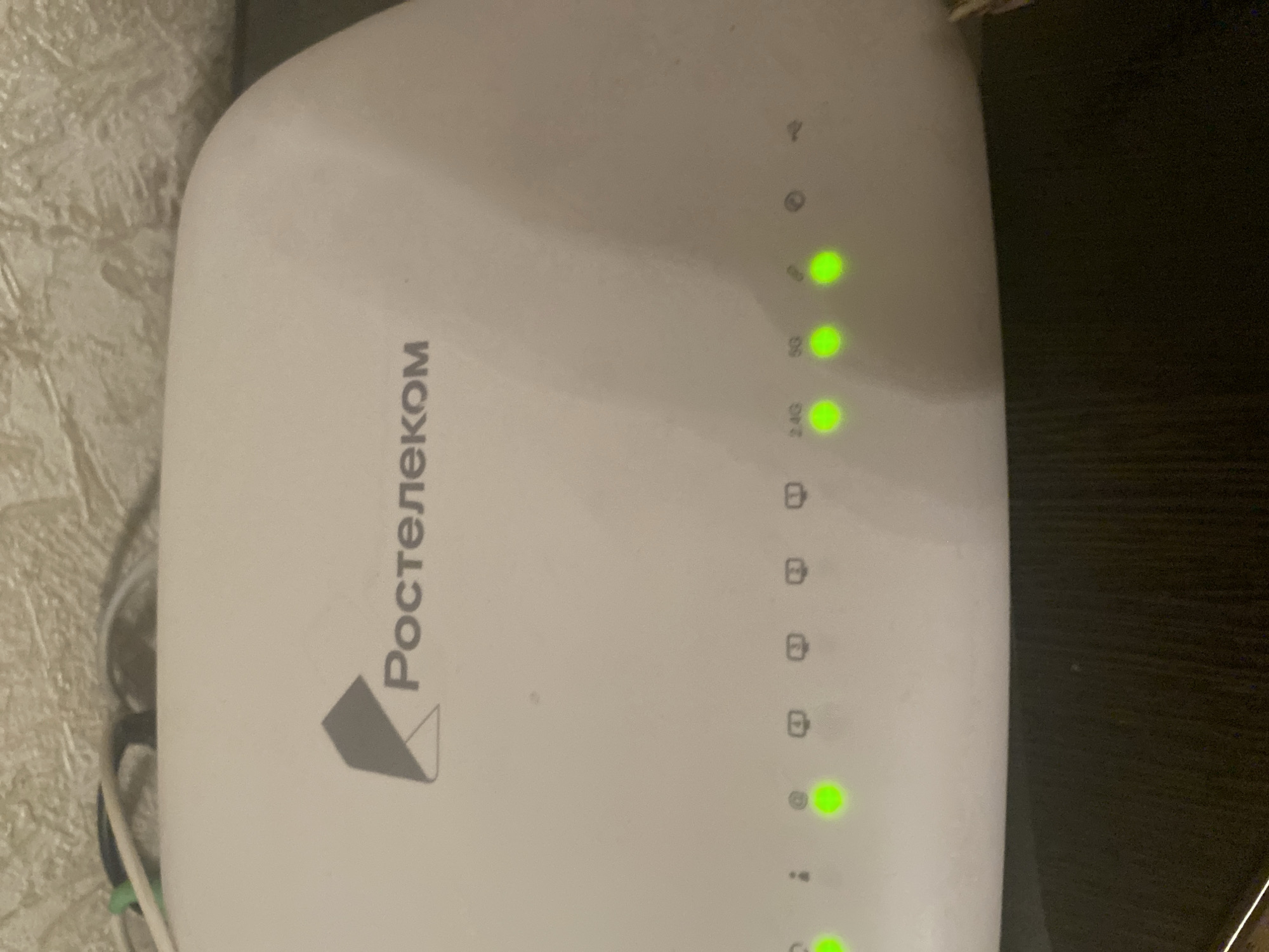 This is an eye-level, close-up shot of the top surface of a white broadband router. The router has the "Rostelecom" logo printed in gray on its top left. Below the logo, a row of LED indicator lights are visible, most of which are illuminated green. From left to right, the visible indicators include what appears to be a power light, an exclamation mark icon, a circle with a dot inside, and symbols that appear to represent Ethernet ports. Labels "2.4G" and "5G" are also visible above two of the green lights, indicating the Wi-Fi bands. The router is situated on a dark surface, possibly a desk or shelf. The background shows a textured wall, likely wallpaper, with some cables visible at the top edge of the router. The overall scene suggests a home or office environment. There is no information about the location in Chernogorsk, Russia.