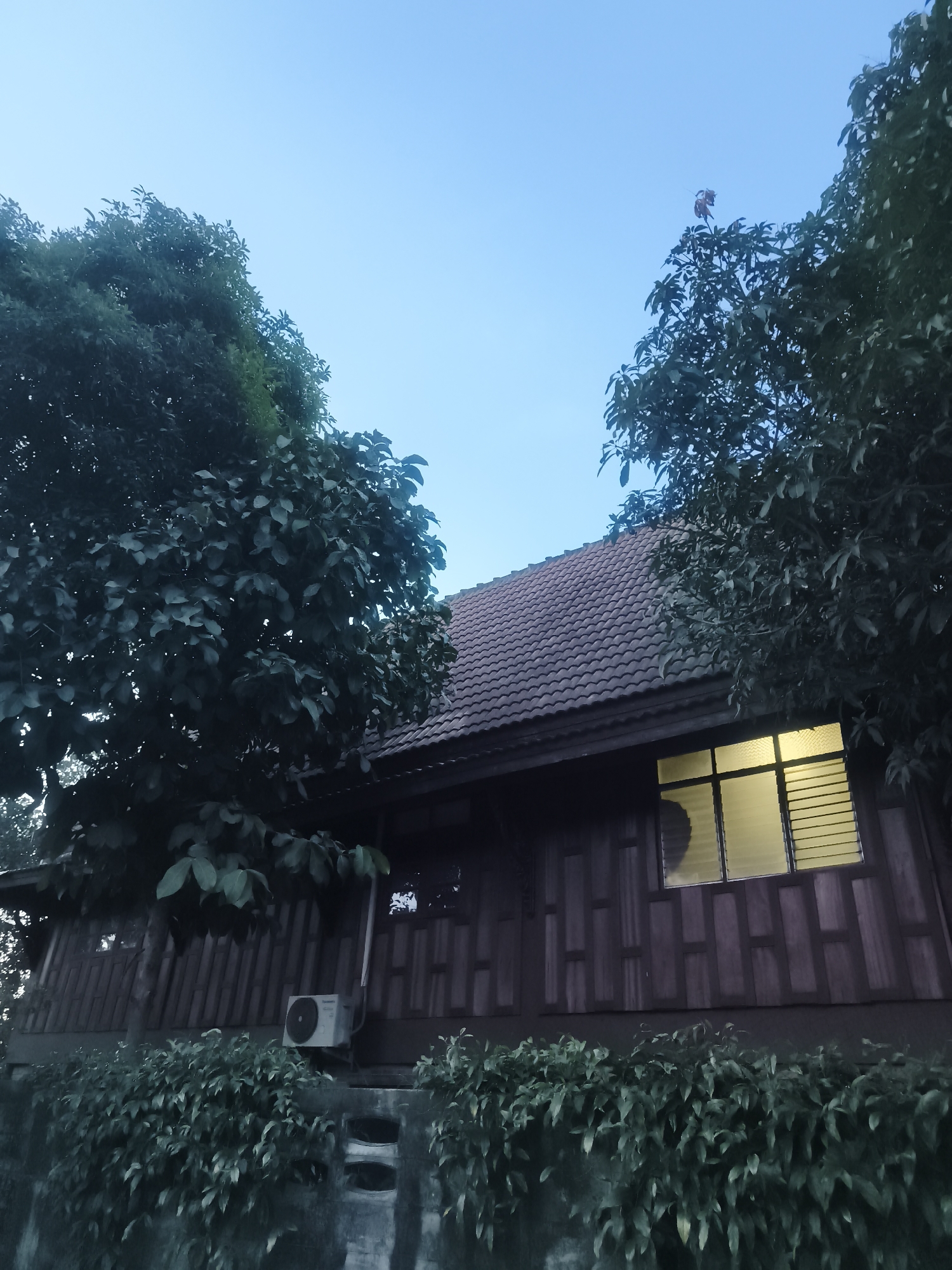 The image depicts a wooden house in Baan Mae Nam, Thailand, likely at dusk or dawn due to the low light and blue sky. The house has a tiled roof and a single visible window with light emanating from within. Trees flank the house, partially obscuring it. An air conditioning unit is mounted on the exterior wall. The house is surrounded by greenery.