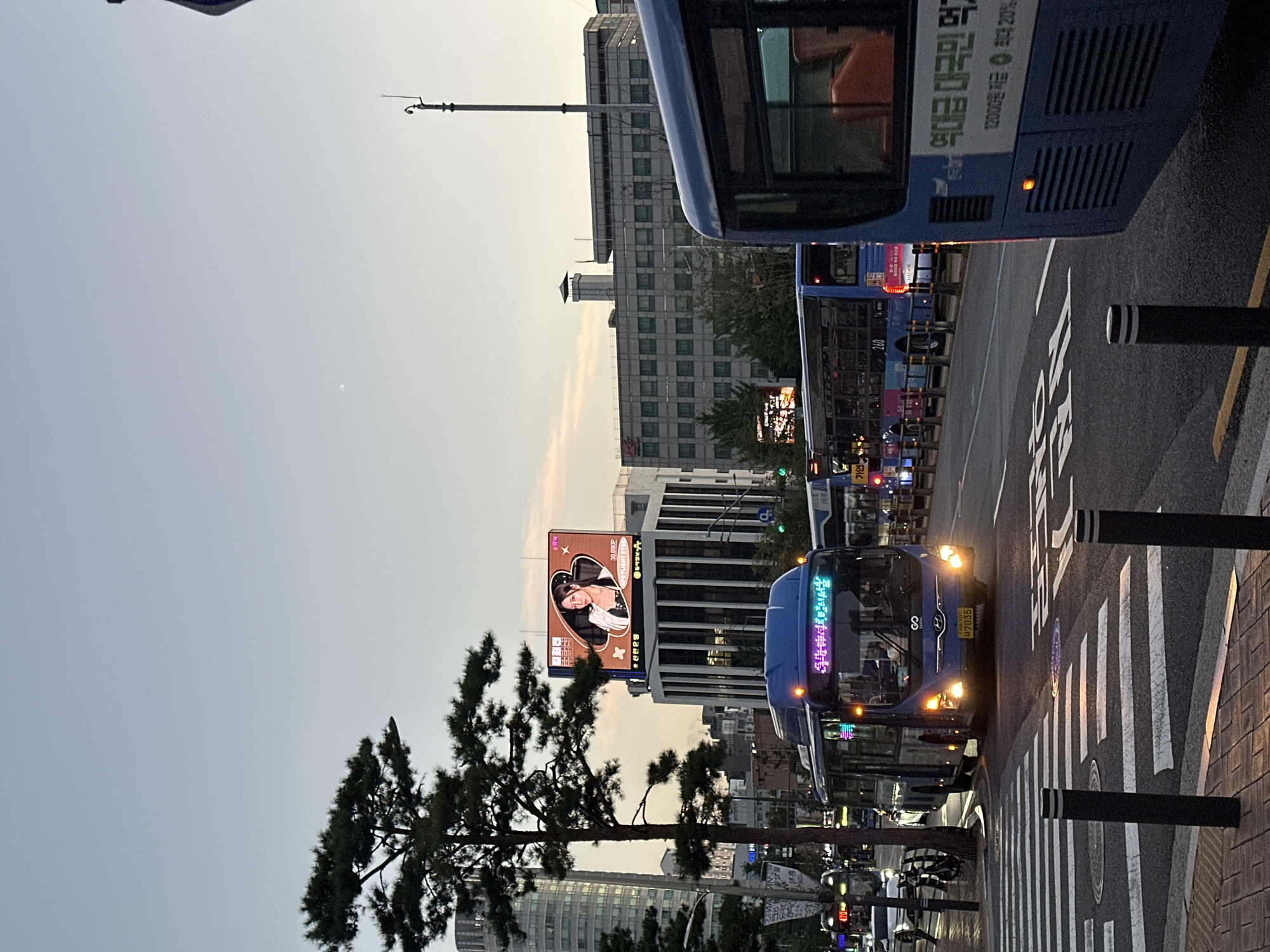 The image depicts a street scene in Seoul, South Korea, likely during the evening or early morning given the soft light and illuminated signs. A blue city bus is in the foreground, stopped at a pedestrian crossing. The bus has its headlights on and displays illuminated text on its front. Another blue bus is visible behind it. In the background, there are several multi-story buildings. One building features a large digital billboard displaying an advertisement with a portrait of a woman. Trees line the street, and streetlights are also visible. The overall impression is a typical urban environment in South Korea.