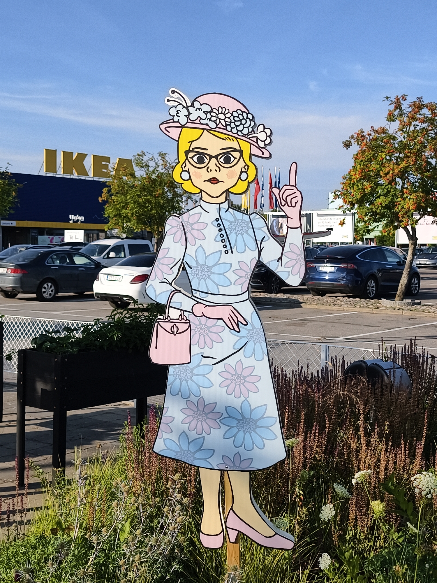 The image depicts a life-sized, cartoon-style cutout of a woman in a hat and a floral dress, standing outdoors. The cutout has blonde hair, glasses, and is wearing a pink hat adorned with flowers and feathers. She is holding a pink handbag and has one finger raised. Behind her, a parking lot is visible, with several cars parked, including a blue Tesla SUV and a white Mercedes sedan. In the background, the yellow IKEA logo is prominent on the blue facade of a building. The scene is set against a clear blue sky with scattered clouds. The cutout is positioned in front of a planter box filled with greenery and surrounded by dry grasses and white flowers. The location is identified as the IKEA store in Linköping, Sweden.