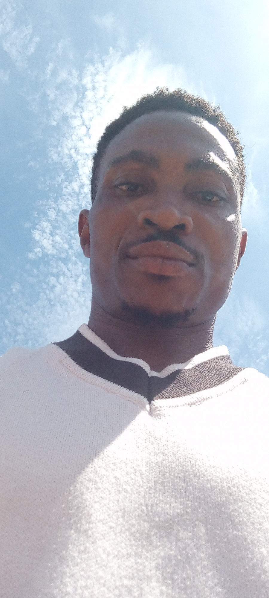 A close-up, low-angle shot features a man with dark skin, a mustache, and short dark hair. He is wearing a white sweater with a dark-colored trim around the collar. The background is a bright blue sky with scattered, fluffy white clouds. This photo was taken in Adankolo, Nigeria.