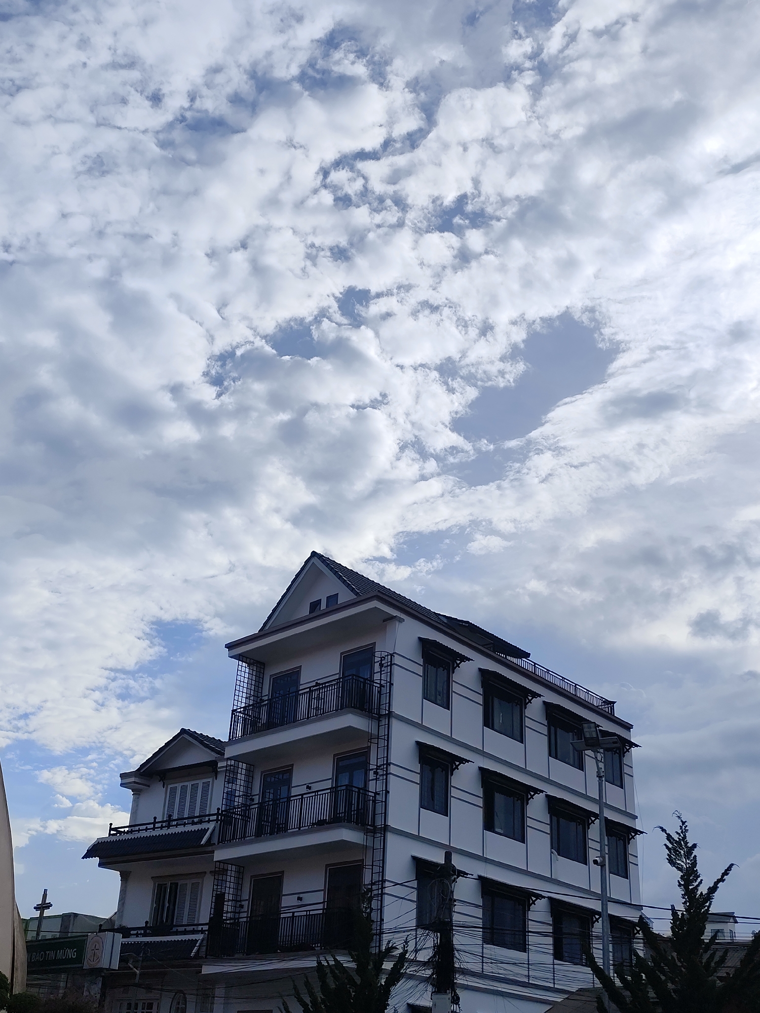 The image shows a multi-story building in Da Lat, Vietnam, under a cloudy sky. The building is primarily white with dark accents on the roof, window awnings, and balcony railings. It has an asymmetrical design with a section featuring gabled roofs and shuttered windows, contrasting with the more modern, flat-roofed section with open balconies. The building's architecture suggests a blend of styles. Below the building, there are trees and some visible power lines. A sign and a cross are partially visible on the left side of the image, hinting at a nearby establishment or religious site. The focus is on the building and the sky above.