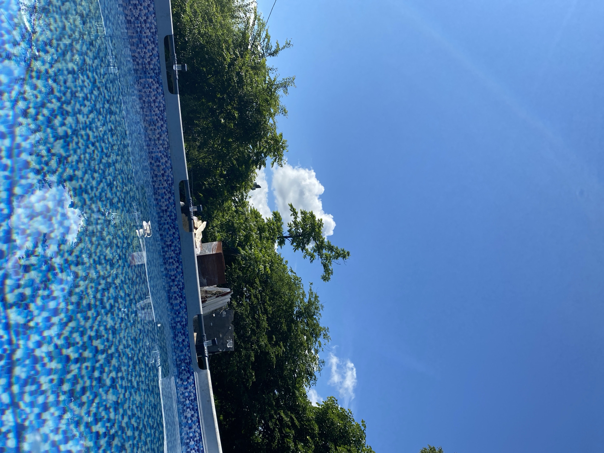 The image depicts an empty swimming pool with a blue tiled interior, reflecting the sky and surrounding trees. The pool's edge, a light gray band with a patterned blue trim, is visible at the bottom of the frame. Beyond the pool, lush green trees form a dense backdrop against a bright blue sky dotted with white clouds. In the upper right corner, a few of the trees are partially obscured by the edge of the frame. The scene appears to be a residential outdoor area. No people are visible in the image. The location is stated to be in Artyom, Russia.