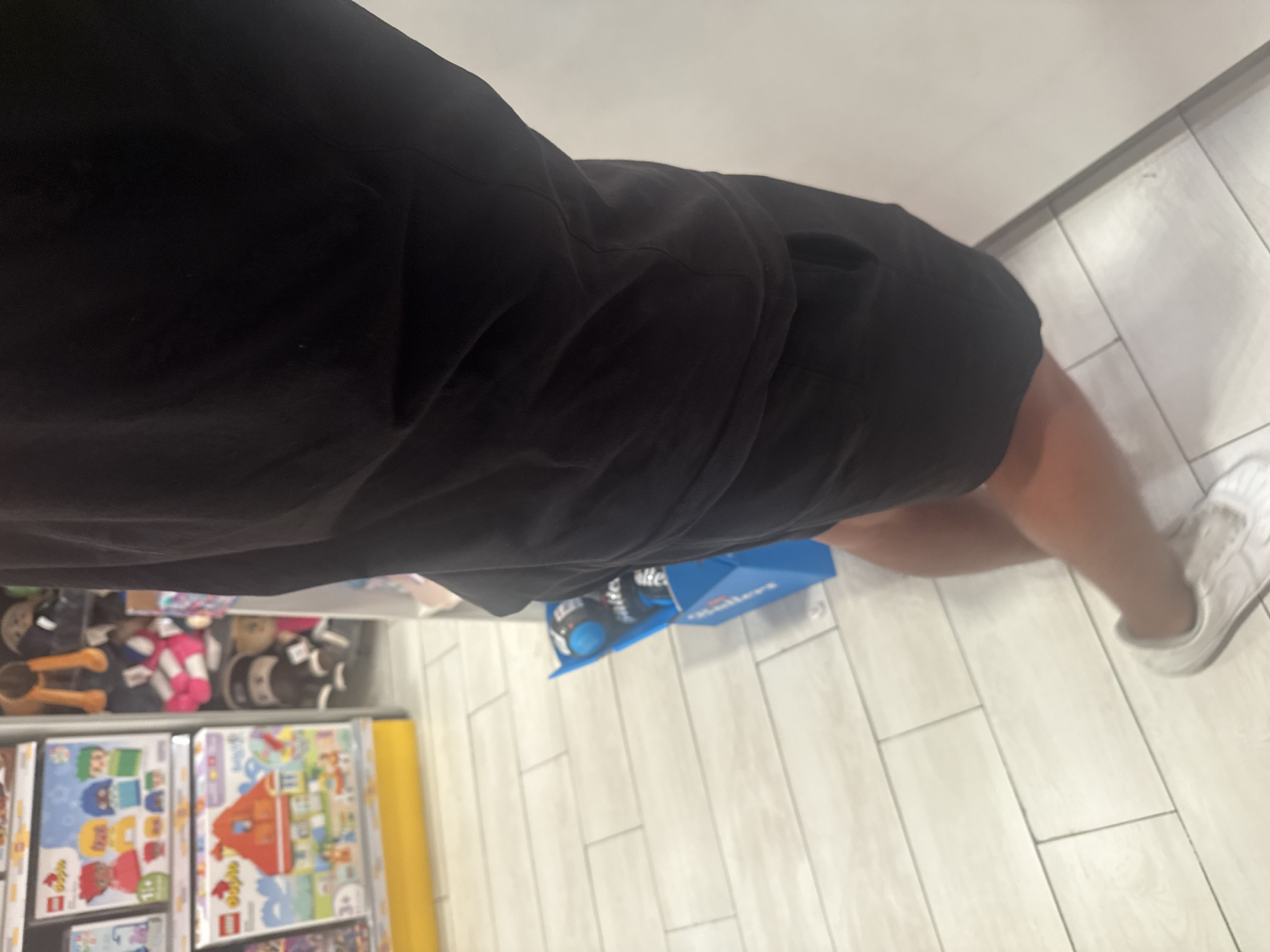 The image shows a person wearing black shorts and white sneakers standing in a store aisle. The aisle contains toys and games on shelves. A blue package sits on the floor near the person's legs. The floor is tiled. The location is presumed to be a store in San Bernardino Tlaxcalancingo, Mexico.