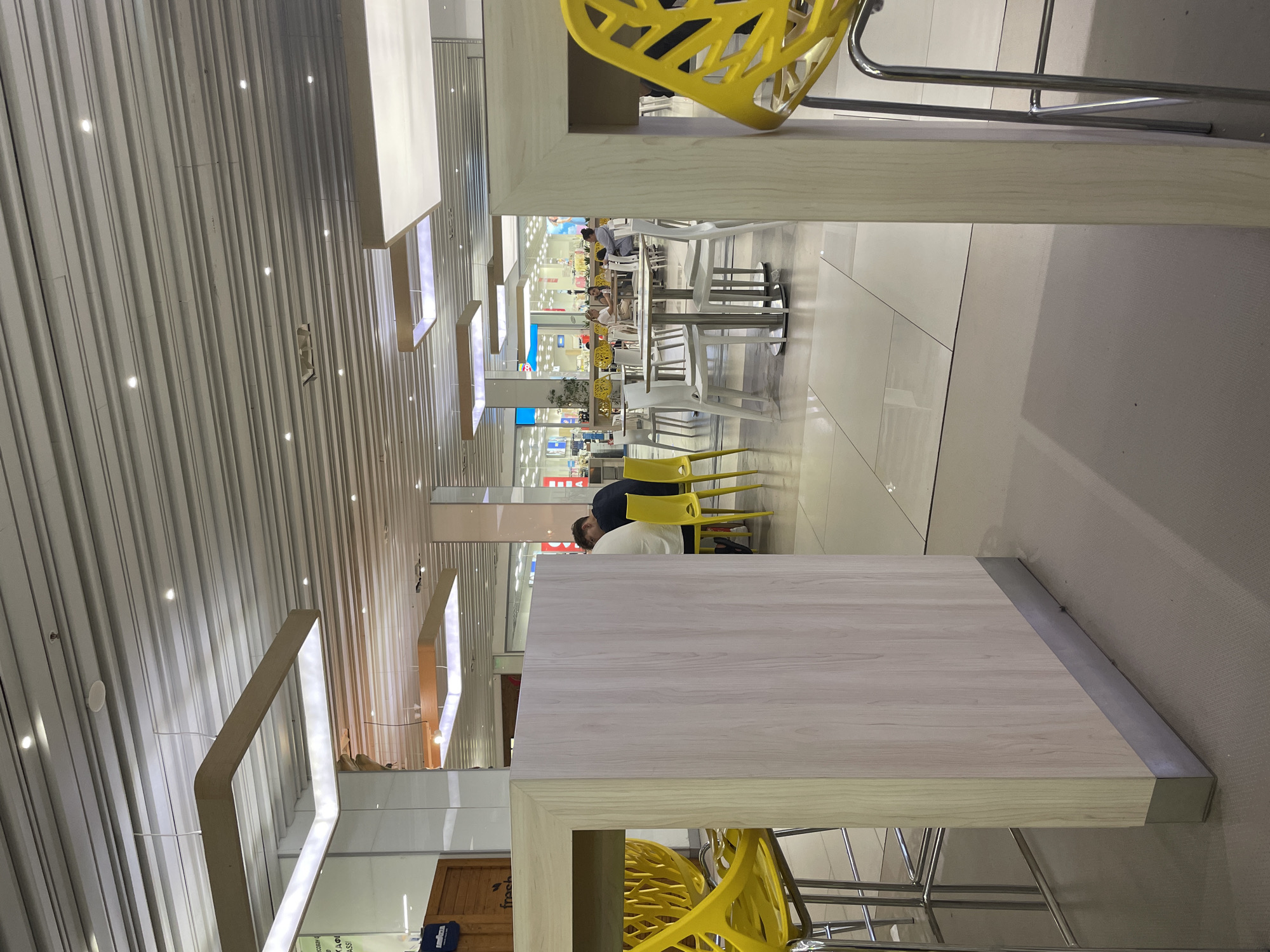 The image depicts a contemporary indoor seating area, likely a food court or cafe, within a commercial establishment. In the foreground, a light-colored wooden partition stands, with bright yellow chairs visible to its left, suggesting a public dining space. Further back, several tables with white chairs are arranged, occupied by a few individuals engaged in conversation or dining. The ceiling features a modern design with recessed lighting and linear elements. The flooring is a tiled surface. The overall ambiance is bright and functional. The location is identified as being in Plovdiv, Bulgaria, though no specific landmarks are visible in the image to confirm this.