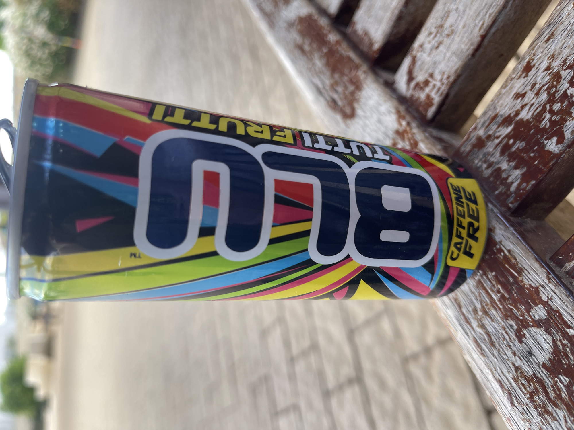 An upright, unopened, full beverage can with a vibrant, multi-colored, abstract design is placed on a weathered, wooden outdoor table, at the bottom of the frame. The can features the word "BLU" in large, white, stylized lettering outlined in dark blue. Above "BLU," the text "TUTTI FRUTTI" is visible in yellow letters. Below "BLU," the words "CAFFEINE FREE" are in yellow text on a dark blue background, also in stylized lettering. The can's top, a silver pull tab, is slightly visible. The background shows a blurred outdoor setting with a paved walkway and foliage, suggesting a casual outdoor location in Even Yehuda, Israel. The table's surface is worn with visible wood grain and peeling paint, indicating age and exposure to the elements. No people are present in the frame.