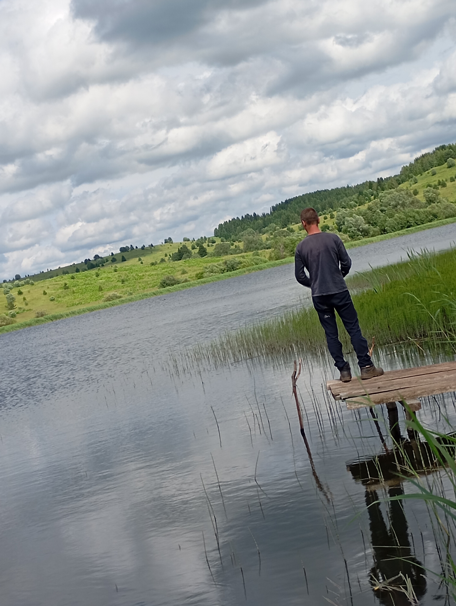 In Ryabki, Russia, a person stands on a wooden pier extending into a calm body of water. The individual, seen from behind, wears a dark long-sleeved shirt and dark pants, with brown shoes. They are positioned facing away from the camera, looking out towards the water and the lush, green landscape beyond. The water's surface reflects the cloudy sky and the surrounding reeds, creating a serene and natural scene. The background features rolling hills covered in dense vegetation and trees, under a sky filled with a mix of white and gray clouds. The overall impression is one of a peaceful, outdoor setting.