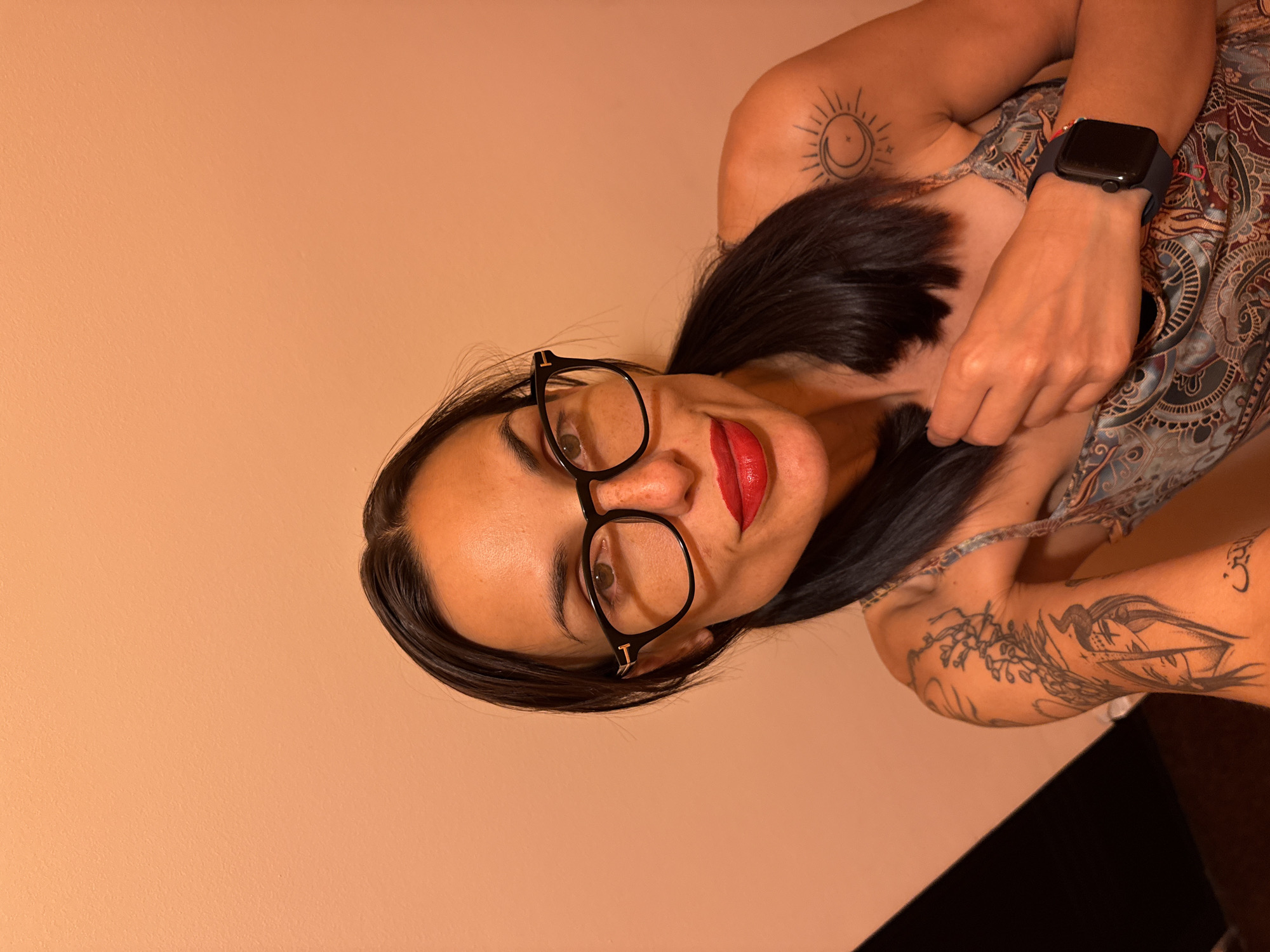 A woman with dark hair and glasses is posing for a selfie. She is wearing red lipstick and has tattoos on her arms and shoulder. She is also wearing a watch on her left wrist. The background is a peach-colored wall. The image does not provide any information about the location in the city of Pleven, Bulgaria.