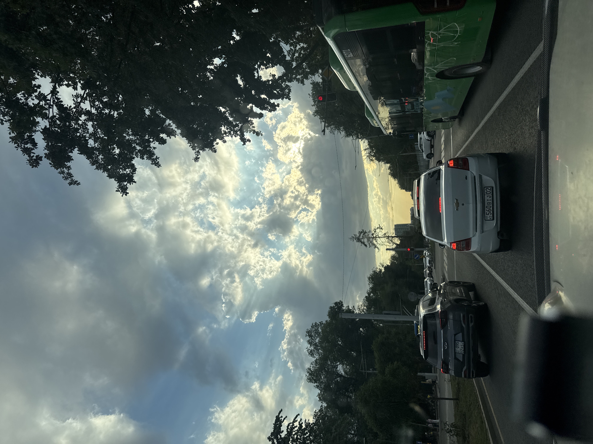A street-level perspective from a vehicle in Almaty, Kazakhstan, captures a traffic scene under a dramatic sky. Sunlight pierces through scattered clouds, creating rays of light that illuminate the sky. In the foreground, the dashboard and windshield of the camera's vehicle are visible. The road ahead shows a line of cars, including a dark SUV and a white sedan. To the right, a green bus with text and images on its side is in motion. Trees line the sides of the road, and a traffic light with a red signal is prominent. In the distance, buildings and more trees form the city's skyline. The scene depicts a typical urban environment during what appears to be late afternoon or early evening, with the interplay of light and shadow adding depth and drama to the cityscape.