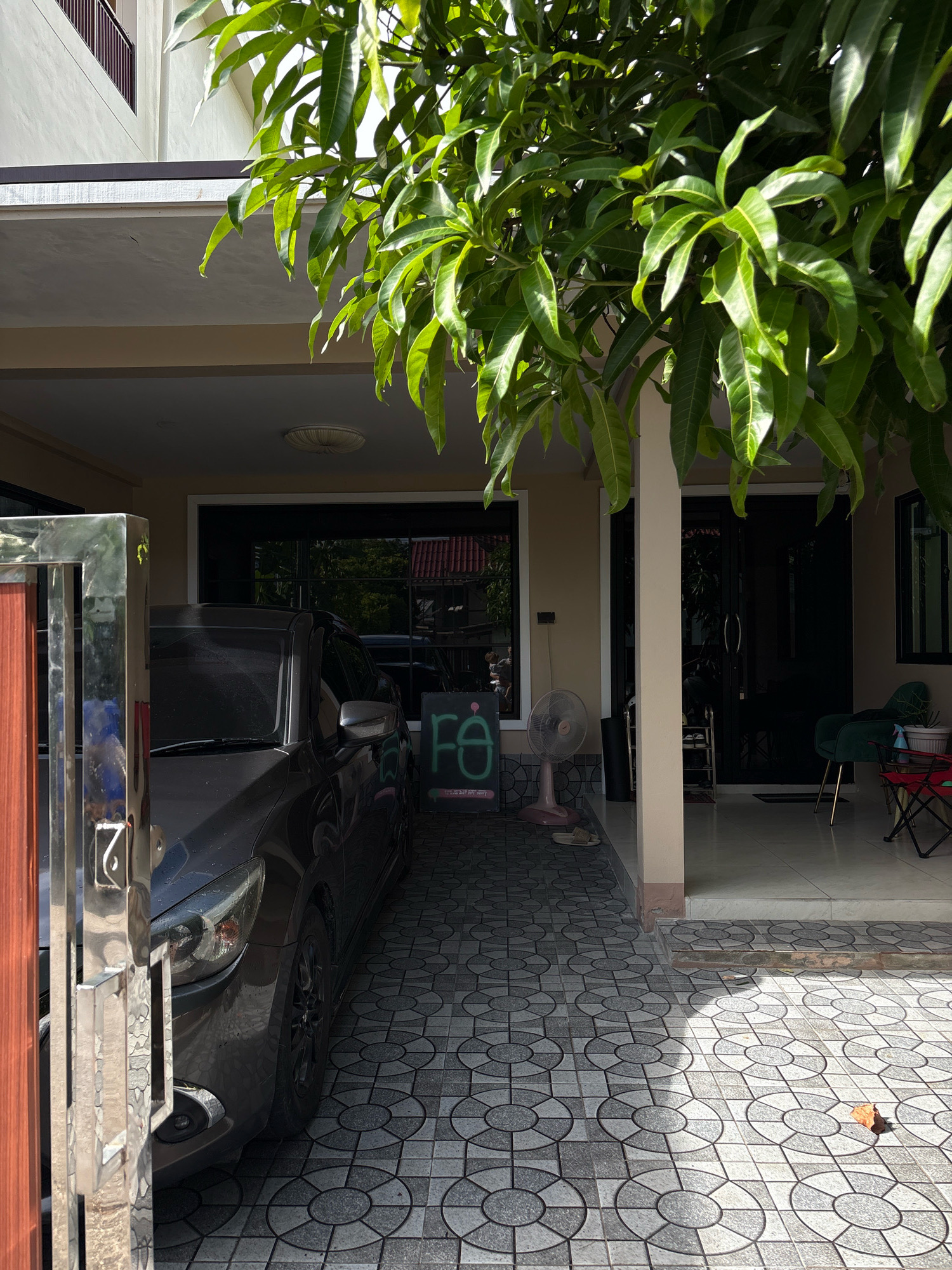 A gray car is parked under a covered carport in front of a building with a beige facade and a dark-tinted glass door. A stainless steel gate with a wooden insert and the number "7" is visible on the left side of the frame. Lush green leaves from a tree overhang the carport. A pink electric fan stands on the patterned tiled ground to the left of the car, near a small chalkboard sign with the letters "fe" in green. The foreground features a gray and white circular patterned tile walkway leading to a raised entrance. To the right, through an open doorway, a dark door with silver handles and a shoe rack can be seen, along with a dark green armchair and a red folding chair. The scene appears to be outdoors, possibly in a residential or commercial area in Choeng Thale, Thailand, with sunlight casting shadows on the ground.