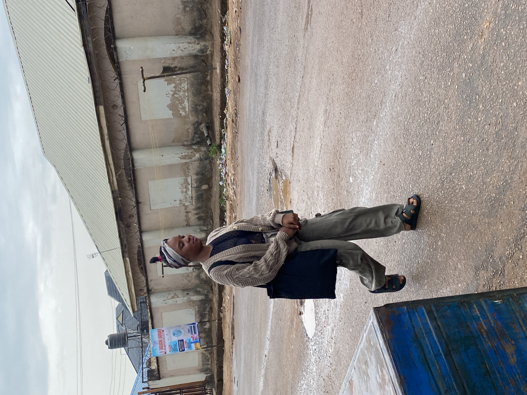 A person is standing on a street in Narayi, Nigeria. The person is wearing a headscarf, a sweater, and pants. They are carrying a dark-colored bag and wearing dark sandals. The street appears wet, and there are buildings on either side. A blue-painted structure is visible in the foreground. A water tank is visible on one of the buildings. A faded poster is fixed to one of the building.