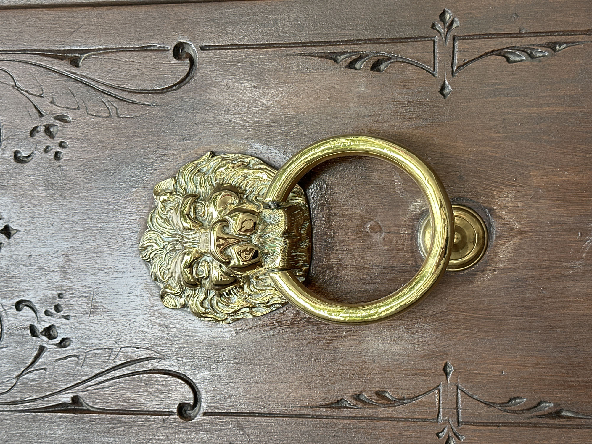 A close-up photograph shows a dark wooden door adorned with a golden lion head door knocker and decorative carvings. The knocker consists of a circular ring held by the lion's mouth. The wood shows signs of wear and age. Based on the architecture style of the door, this is likely to be located somewhere in the city of Madrid, Spain.