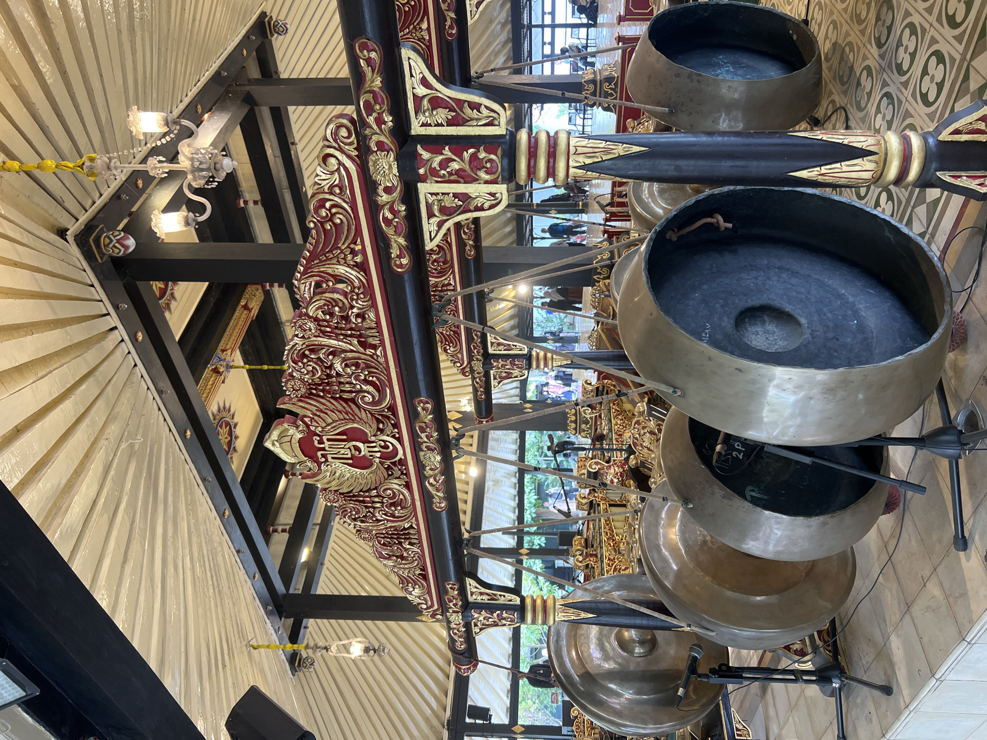 A wide shot captures a Gamelan ensemble, a traditional Indonesian orchestra, within a Pavilion in Yogyakarta, Indonesia. The Gamelan instruments, primarily large bronze gongs, dominate the foreground and midground. They are suspended from ornate wooden frames decorated with red and gold intricate carvings and motifs. The instruments themselves are made of polished bronze and vary in size, some with visible hammered patterns. Microphones are attached to several of the gongs, suggesting a live performance or recording.
Behind the instruments, the pavilion features a high, sloped ceiling with a fan-like design of pale wood beams. Black wooden pillars support the structure, and decorative elements in red and gold are visible on the ceiling and the frame of the instruments. Chandeliers with multiple lights hang from the ceiling, contributing to the warm, ambient lighting.
In the background, a number of people are visible, appearing to be spectators or participants, scattered around the pavilion. Some are seated, others are standing and observing. The setting appears to be an open-air or semi-open-air structure, with glimpses of greenery and daylight through the openings. The floor is tiled with a patterned design. The overall impression is one of a cultural performance space, showcasing the rich artistry of Indonesian music and architecture.