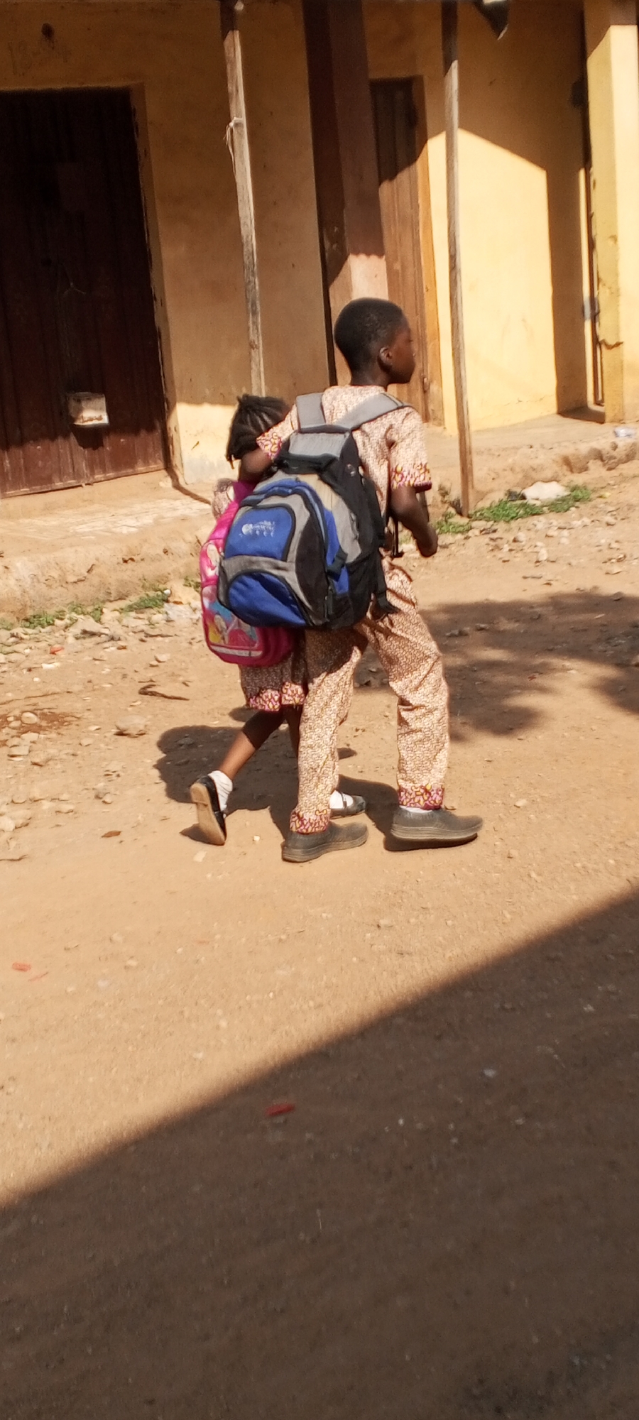 The image depicts two children on a dirt path in Sabon Tasha, Nigeria. The older child carries a larger backpack while the younger child carries a smaller, pink backpack. Both children are wearing patterned clothing. The setting appears to be a residential area with buildings visible in the background. The area is bathed in sunlight.