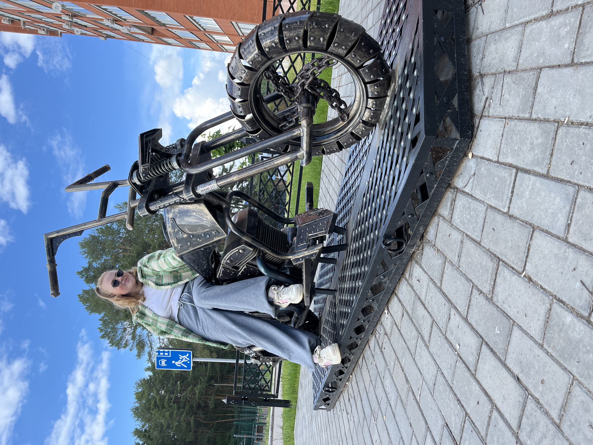 An outdoor, eye-level shot depicts a young woman casually seated on a large, custom-built metal motorcycle sculpture on a bright, sunny day. The image is taken in a public area, with a modern brick apartment building visible on the left and a backdrop of blue sky with scattered white clouds and green pine trees.
The woman, with blonde hair, wears sunglasses, a white t-shirt, a green and white plaid overshirt, and grey sweatpants paired with white and pink athletic shoes. She is positioned comfortably on the sculpture, with her left arm resting on the handlebar, looking towards the camera with a relaxed expression.
The motorcycle sculpture is predominantly black and metallic grey, featuring intricate details crafted from metal. Its front wheel is distinctive, made of multiple metal segments, and the hub incorporates chains. The entire sculpture is securely mounted on a robust, textured black metal platform. Visible on a part of the motorcycle frame are two labels with Cyrillic text: "КАСТОМ" (Custom) and "МОНКИ.777" (Monkey.777).
In the background, a blue road sign with white pictograms, indicating a bus stop and a pedestrian area, stands near the trees. The ground beneath the sculpture and extending into the foreground is paved with grey rectangular stones. The scene suggests a relaxed moment, possibly a photo opportunity with public art in Chaikovsky, Russia.
