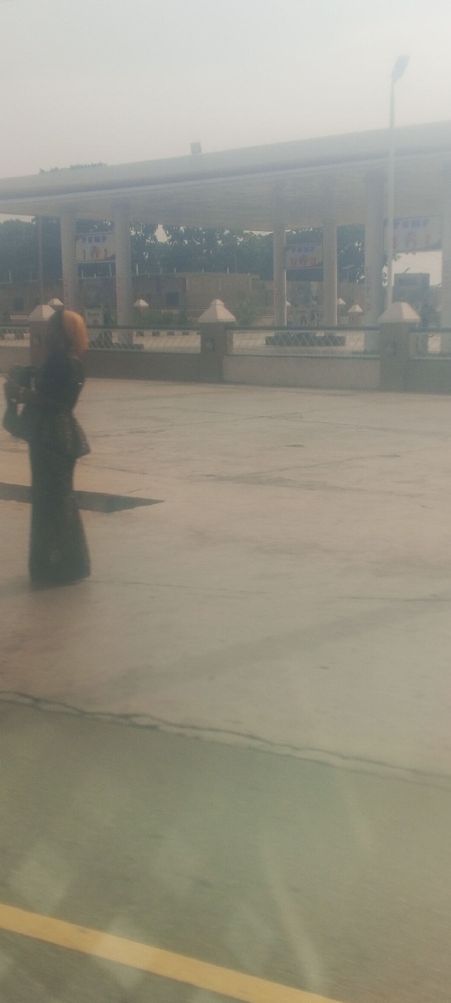 The image shows an individual standing in front of a building in Barnawa, Nigeria. The person is dressed in black, and the building appears to be a commercial or public structure with columns and a covered area. The foreground is a paved or concrete surface, and the overall lighting suggests it is daytime.