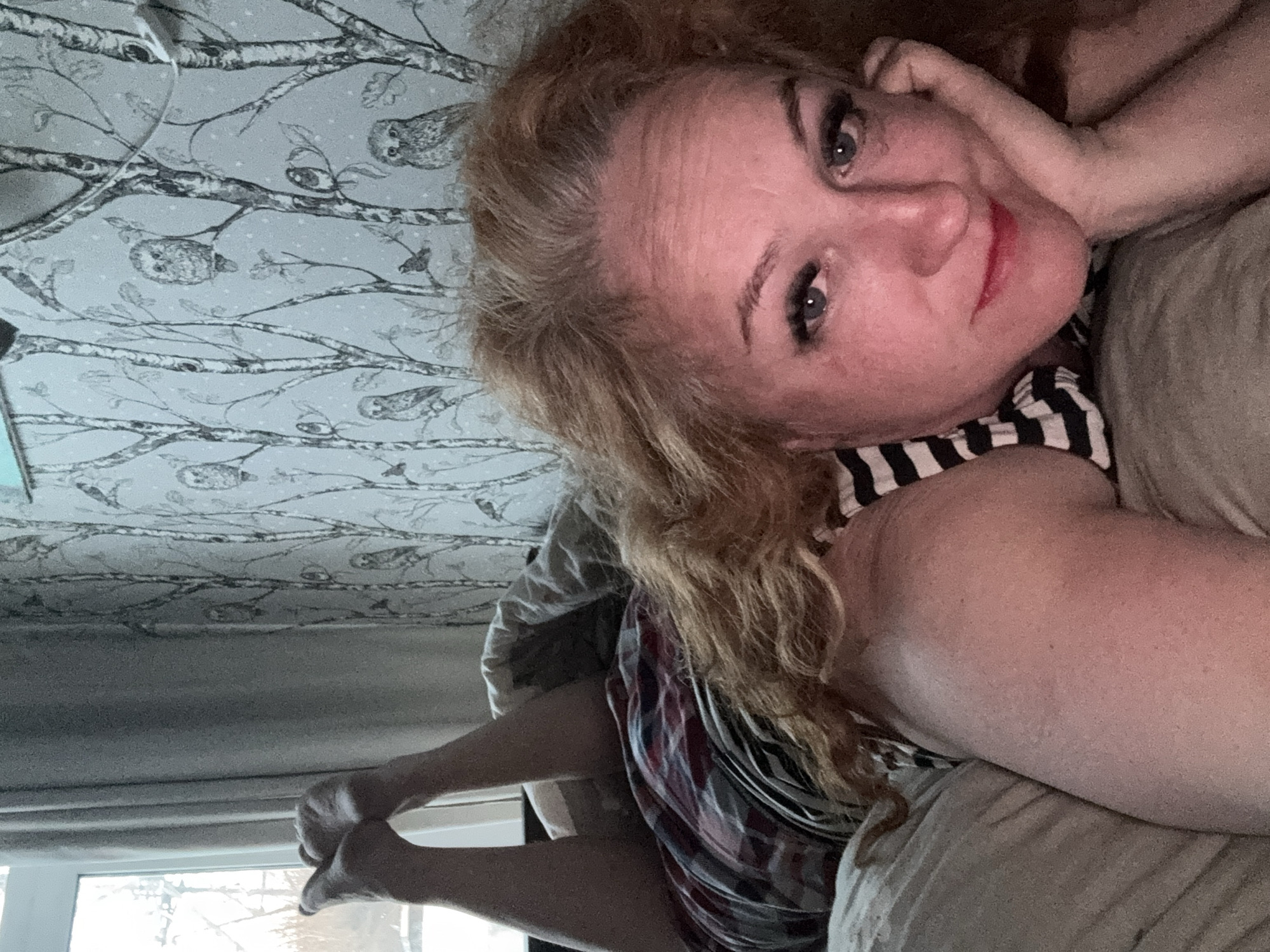 The media file presents an upward-angled selfie of a woman with reddish-blonde, curly hair and blue eyes, captured in a relaxed indoor setting. She is lying down, likely on a bed or sofa, with her head resting comfortably on a plain, light-colored cushion. Her expression is serene, featuring a subtle smile and a hint of red lipstick.
She is dressed in a black and white striped sleeveless top and plaid shorts with red, white, black, and blue patterns. Her bare feet are visible in the lower left portion of the image, appearing to be raised towards a window. Through the window, an outdoor scene with bright, diffused light suggests an overcast day or possibly a snowy environment, aligning with the Chaikovsky, Russia context.
The background behind and above her is adorned with light grey or blue wallpaper featuring a distinctive pattern of dark, thin tree branches, resembling birch trees, and numerous small, stylized owls perched among the foliage. The overall atmosphere conveys a sense of comfort and repose. No text is visible in the image.