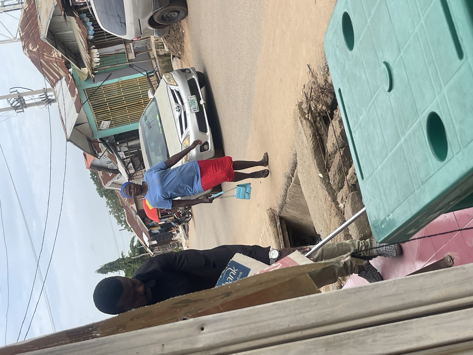 In Abuja, Nigeria, a person is walking on a dirt road with a purse in their hand, wearing a blue shirt and a red skirt. A silver car is parked on the side of the road. Another person is standing behind them, wearing a black shirt and khaki pants, holding a box with the logo of a bank. In the foreground, a green plastic table is visible. Buildings with rusty roofs line the street, and electrical wires are strung overhead.
