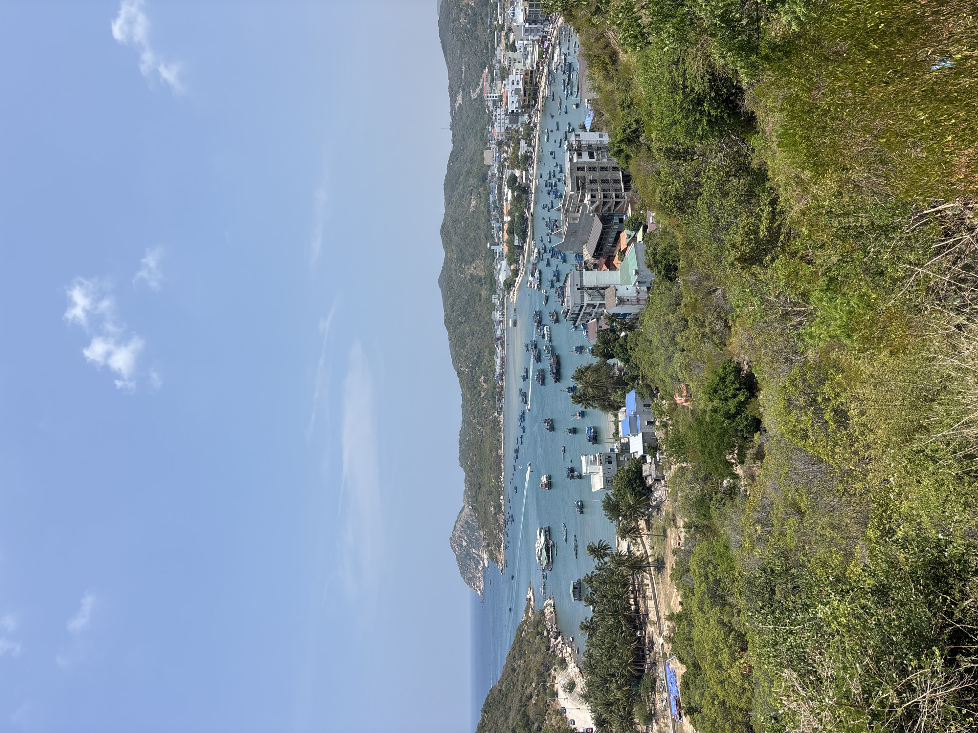 The image shows a view of a bay in Vĩnh Hải, Vietnam from an elevated position. The bay is filled with numerous boats, with the town built along the coastline. Lush green vegetation covers the foreground on the right side of the image and is also visible on the hills in the background. The sky is a clear blue with a few scattered clouds. Buildings and infrastructure are present along the shoreline.