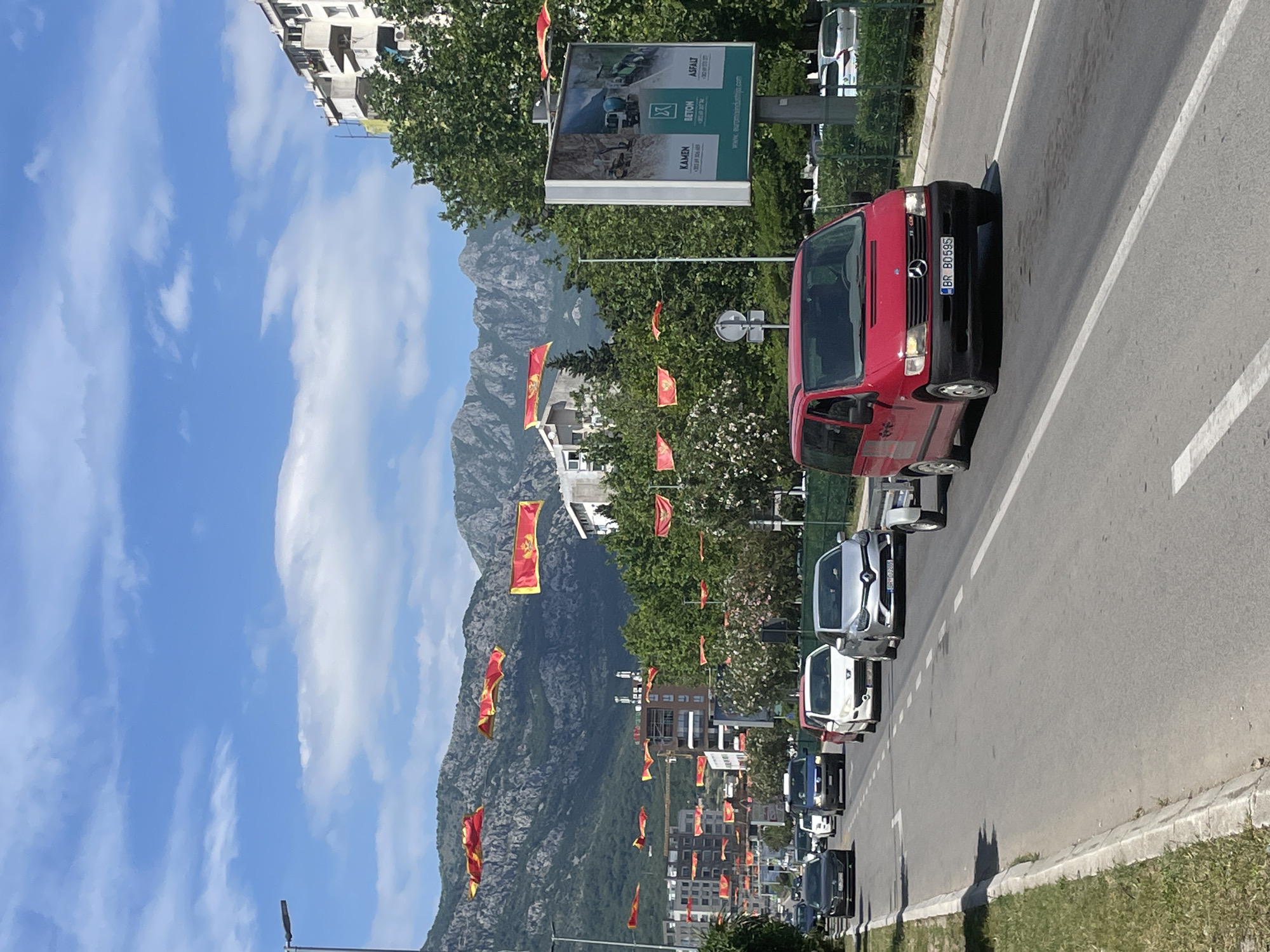 The image shows a street scene in Bar, Montenegro. A row of cars, including a red Mercedes van with license plate "BR BD595", are parked along the right side of the road. Numerous Montenegrin flags are displayed on poles lining the street. In the background, a large mountain rises prominently. A billboard is visible on the right, displaying advertising. The sky is blue with scattered clouds. Buildings can be seen in the background, along with trees. The scene is bathed in bright daylight.