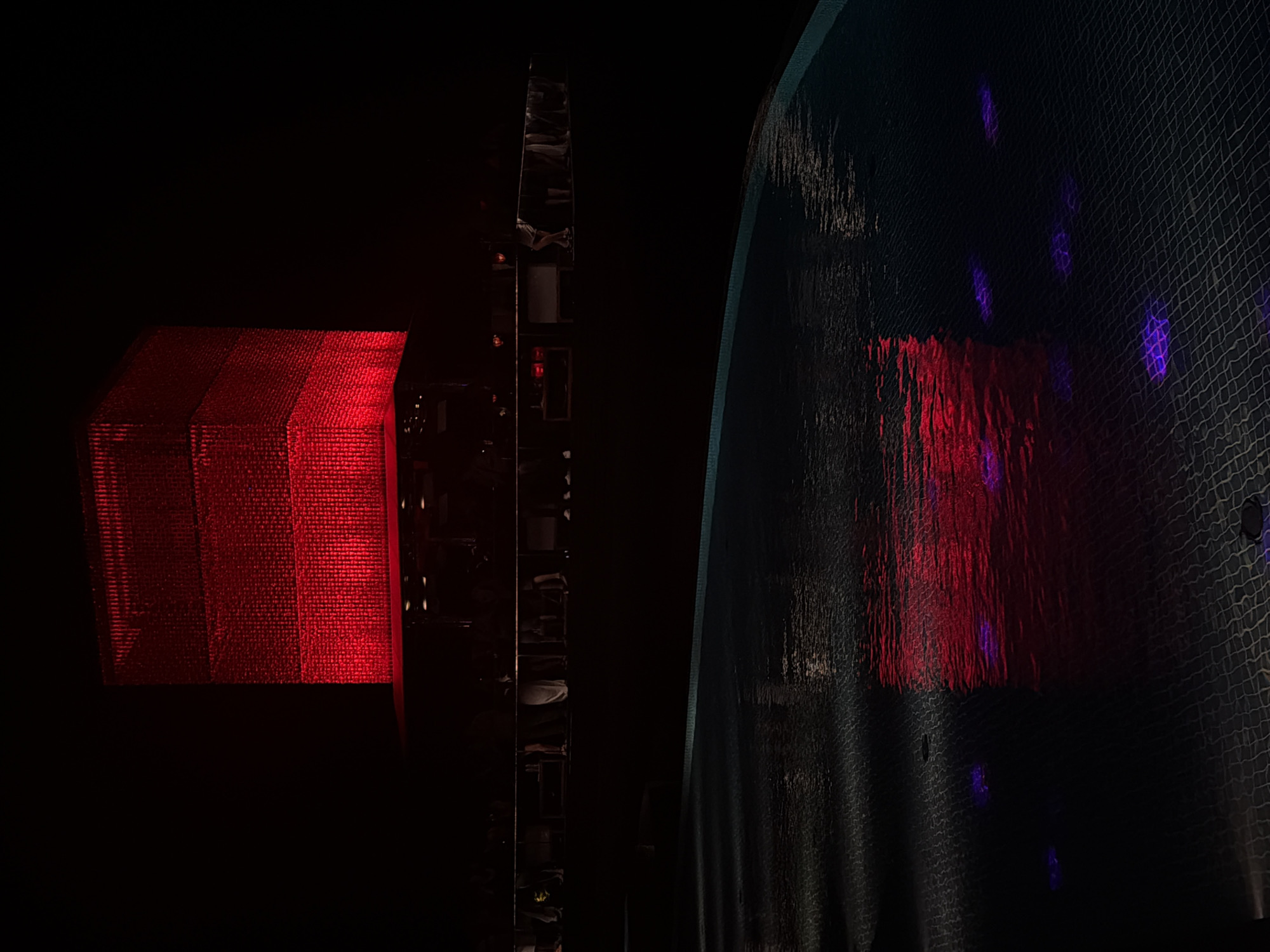 Captured in Kuta Selatan, Indonesia, the image features a dimly lit, primarily nocturnal setting. A cubic structure illuminated with red light dominates the left side of the frame, revealing a textured, possibly metallic or reflective surface. Adjacent to it, a dark, narrow structure with visible shelves and objects sits vertically. To the right, a curved wall or surface with a grid-like pattern is partially illuminated by what appears to be a reflection of the red light, intermixed with additional, scattered purple lights. The overall ambiance is dark, with the scene's elements defined by artificial lighting against a black background.