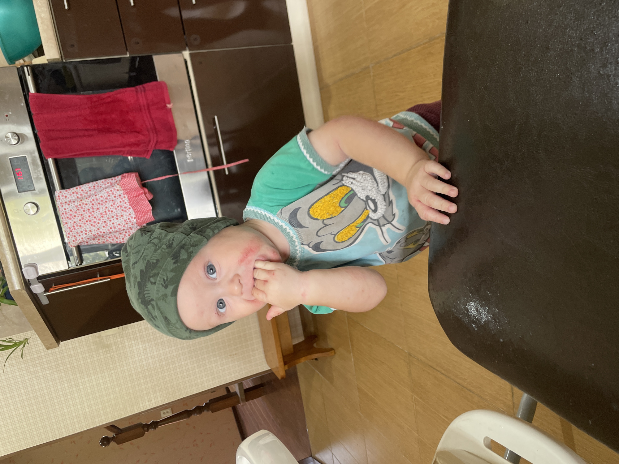 A young child wearing a green dinosaur-patterned beanie and a t-shirt with a cartoon character on it is standing in a kitchen. The child has red smudges around their mouth and is putting their fingers in their mouth. In the background, there is a stainless steel oven with red and white towels hanging on it, dark brown cabinets, and a wooden floor. A wooden stool is visible to the left of the child, and a white high chair is partially visible in the foreground. The scene appears to be indoors, likely in a residential kitchen.