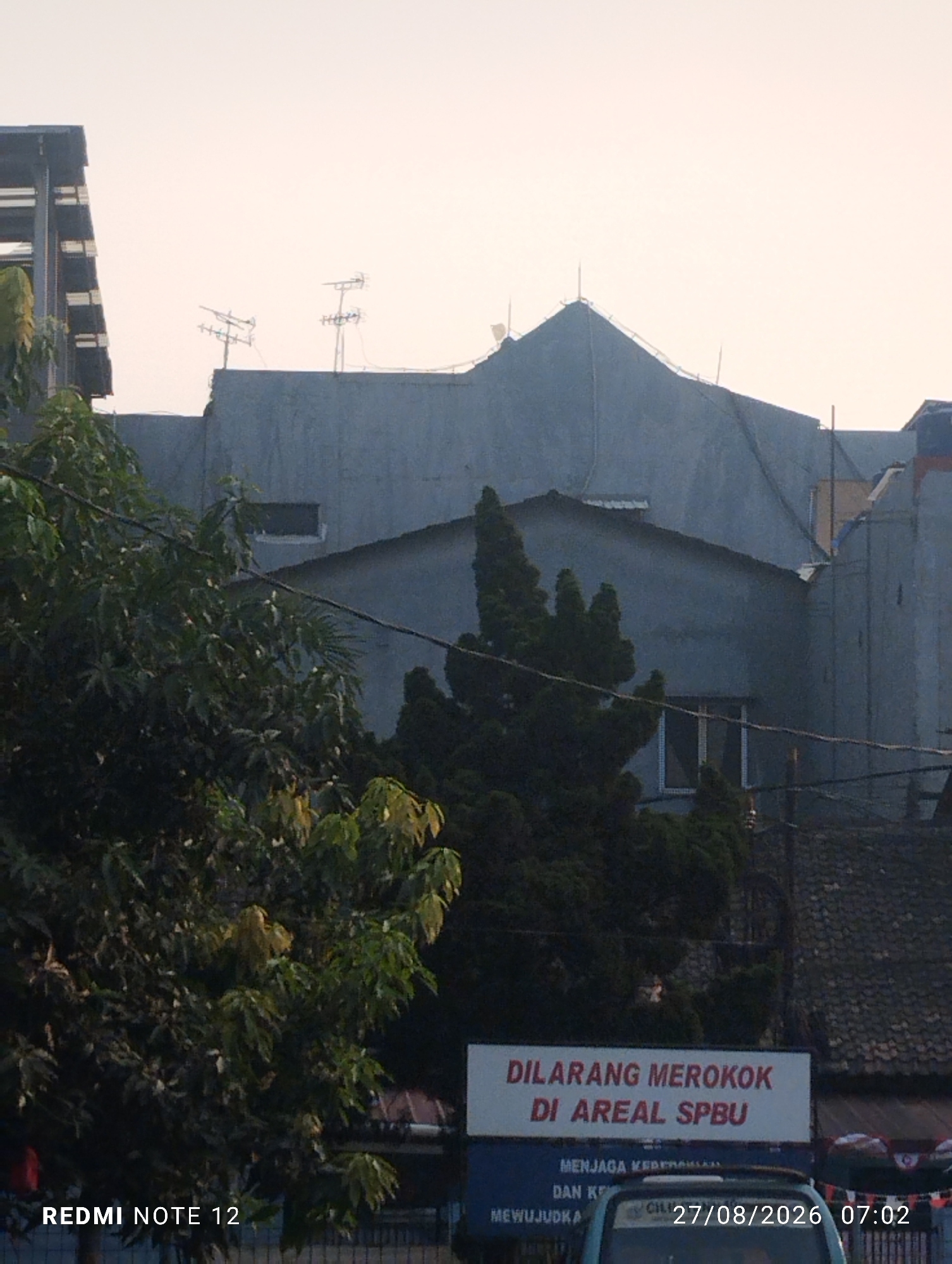 The image, taken in Bekasi, Indonesia, captures a street scene dominated by buildings and foliage. In the foreground, lush green trees partially obscure the view of a building with a blue sign that reads "DILARANG MEROKOK DI AREAL SPBU," indicating a no-smoking zone at a gas station. Below this sign, another blue sign is partially visible. A vehicle, possibly a van, is parked behind the signs, and a timestamp "27/08/2026 07:02" is superimposed on the image. The background features a multi-story building with a grey facade and visible windows. Rooftop antennas and electrical wires are also present, suggesting an urban environment. The sky is overcast, casting a muted light on the scene. The text "REDMI NOTE 12" is also present in the bottom left corner, likely indicating the device used to capture the image.