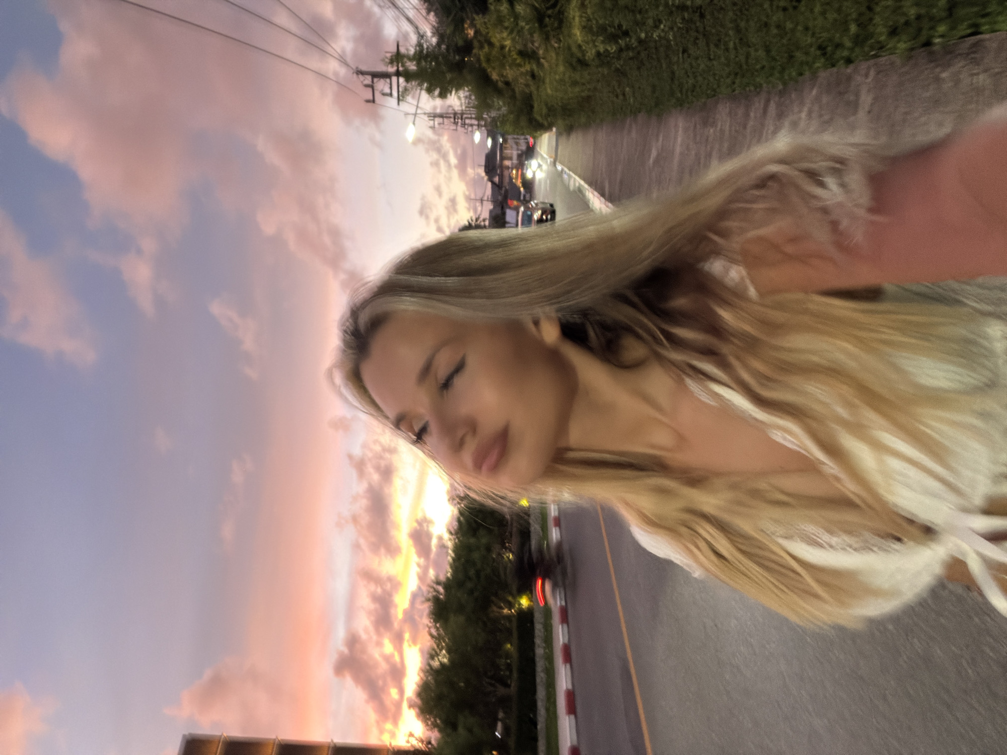 The media file shows a young Caucasian woman with long blonde hair, taking a selfie outdoors. She is facing left with her eyes closed and lips slightly parted, appearing relaxed. She is wearing a light-colored top with a tie detail. Her hair is windswept, suggesting movement.
The background depicts a street scene at sunset. The sky is filled with soft pink and orange clouds, indicating dusk. There are power lines visible against the sky, and trees line the side of the road. A road with painted lines runs through the scene, with a car in the distance and a motorcycle in motion, its taillights blurred, suggesting traffic. The setting appears to be a tropical location, possibly Thailand given the provided location context of Choeng Thale. The overall mood is serene and perhaps a bit dreamy due to the sunset colors and the subject's expression. There is no visible text in the media file.