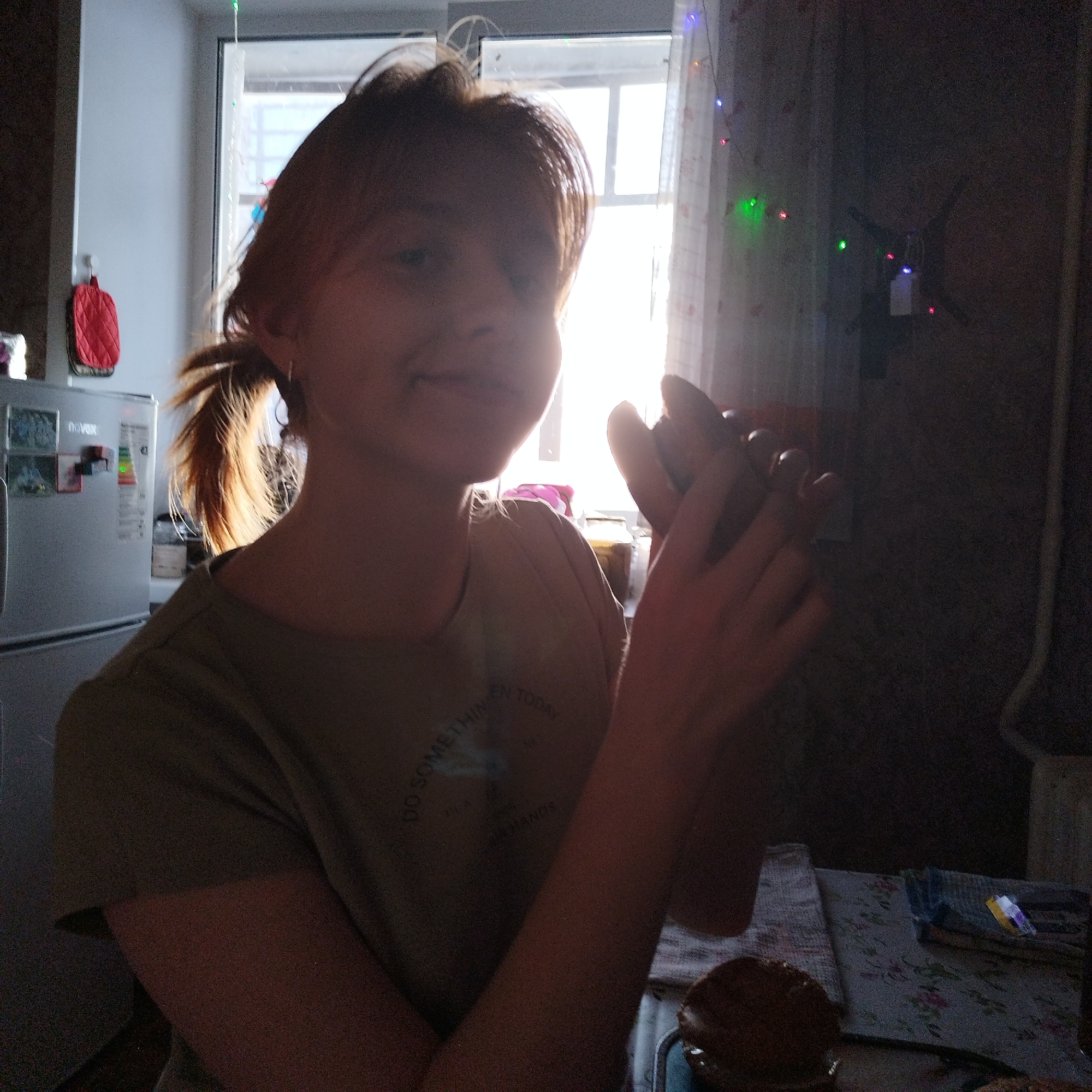 A person is captured indoors, likely in a kitchen or dining area, bathed in strong natural light streaming through a window directly behind them. The individual, with light brown hair tied back in a ponytail, is seen from the chest up, with their face partially obscured by the bright backlight. They are wearing a light-colored (possibly olive green or beige) t-shirt, on which partially visible text reads "DO SOMETHIN EN TODAY" and "YOUR HANDS".
The person's hands are clasped together in front of them, appearing to be actively shaping or working with something, possibly dough or another food item. To their left, a silver refrigerator is visible, adorned with magnets and an energy efficiency label. A red oven mitt or potholder hangs on the wall nearby. Behind the person, a large window is the primary light source, featuring sheer curtains and a string of colorful fairy lights (green, red, blue) draped across it. In the lower right corner, a table with a floral-patterned tablecloth holds a dark, round food item, resembling a bun or bread, on a tray or plate. The overall impression is one of a daytime setting, focused on a domestic activity like baking or cooking. The person has a slight smile, suggesting engagement and perhaps contentment.