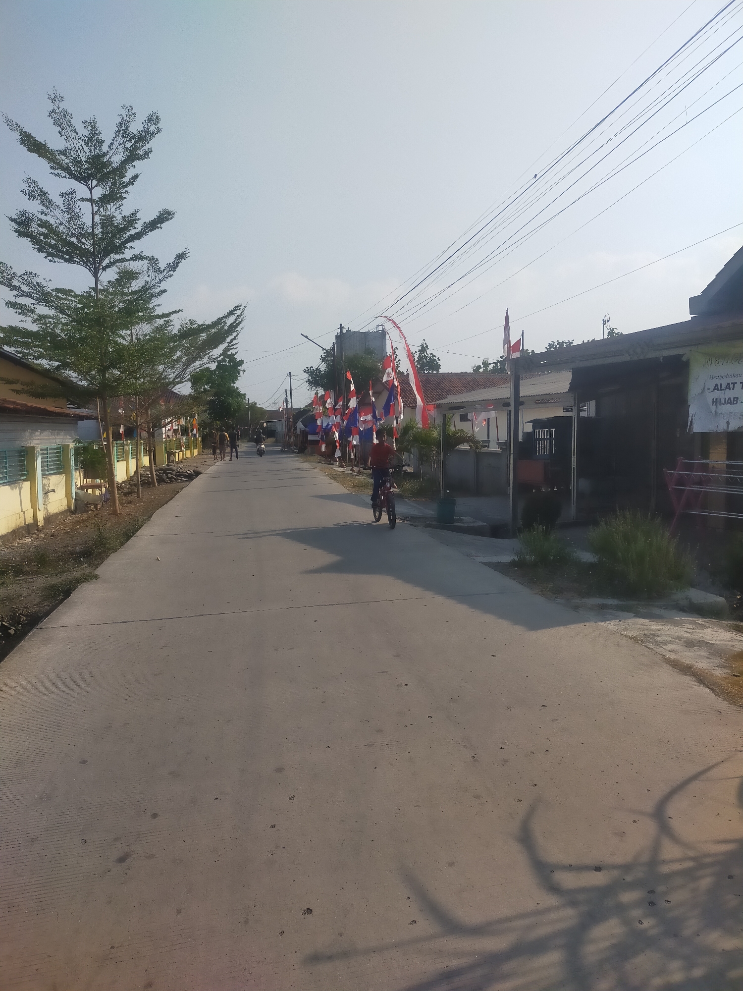 The image depicts a street scene in Sumingkir, Indonesia, likely during a festive occasion, as evidenced by numerous red and white Indonesian flags lining both sides of the road. The street is paved and appears to be a residential or small commercial area, with houses and some shops visible. The sky is bright and clear, suggesting a sunny day.
In the foreground, a young person is riding a bicycle on the street. They are wearing a red shirt and blue pants. Further down the street, a few people are walking, and a motorcycle is also visible. The left side of the street features a row of yellow-painted buildings with green window frames, interspersed with trees and vegetation. The right side of the street has a dark-colored building with a sign that appears to advertise "ALAT T... HIJAB...". Shadow patterns from trees and possibly overhead wires are cast on the road. The overall impression is of a typical Indonesian village or small town street.