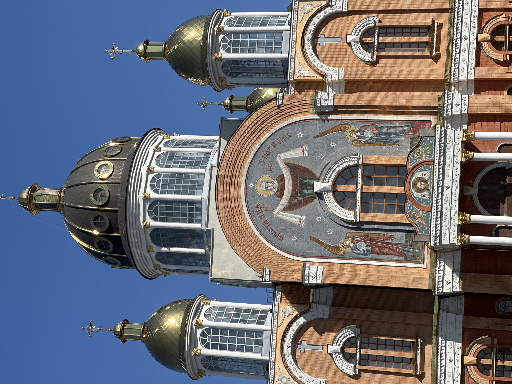 The image captures the architectural details of a church located in Kyiv, Ukraine. Visible elements include brick walls, golden domes topped with crosses, arched windows, and religious iconography. The mosaics depict images of angels and a central figure, likely a saint or Jesus. The architecture suggests an Orthodox Christian church. The sky is clear and blue, indicating fair weather conditions.