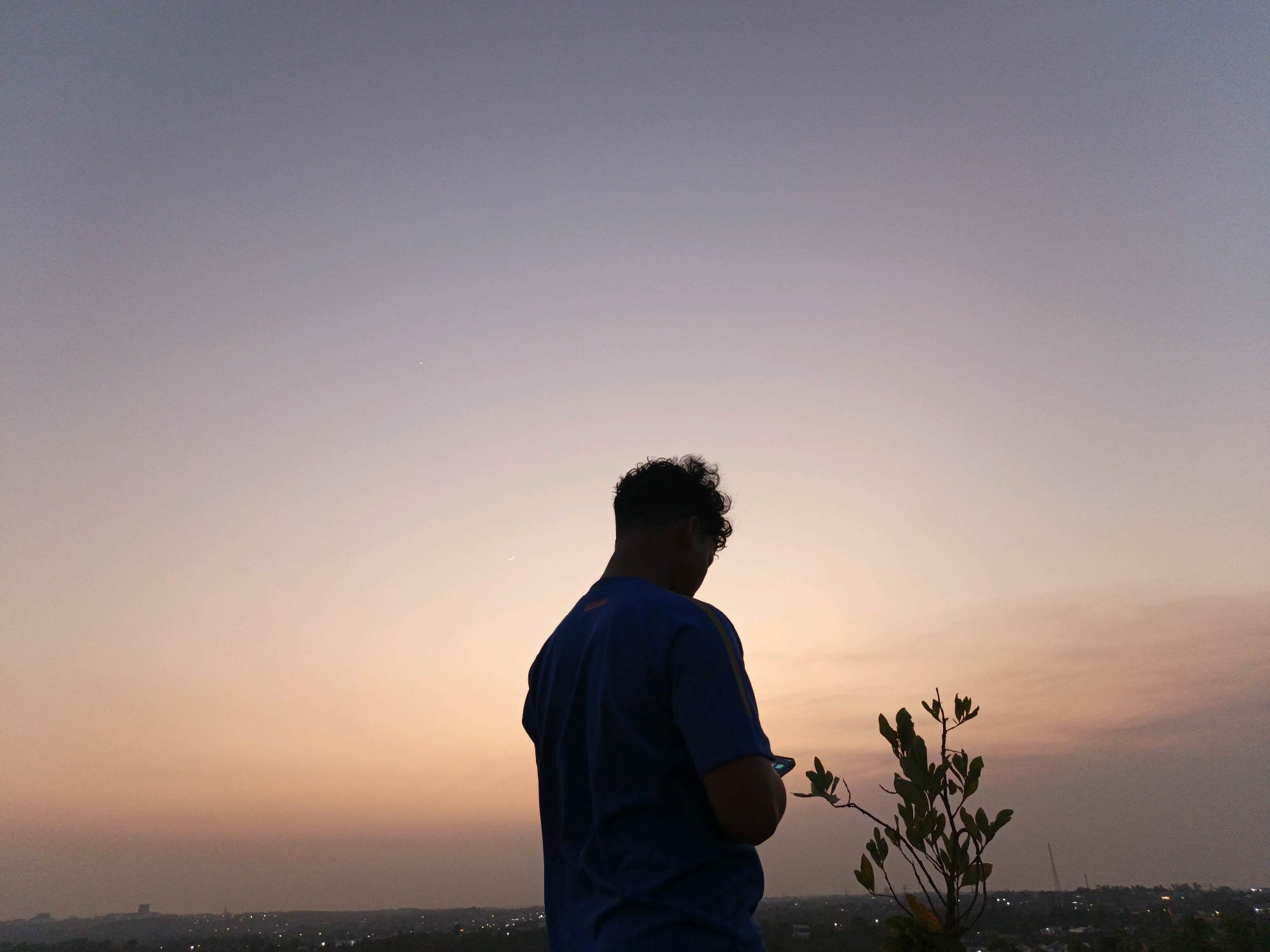 A silhouette of a man in a blue t-shirt stands on a high vantage point, looking at his phone. He is positioned on the right side of the frame, with a small, leafy plant to his right. The background is a gradient of colors, transitioning from a soft purple at the top to a warm orange near the horizon, indicating either a sunrise or sunset. Below the horizon, the lights of the city of Banjarbaru, Indonesia, are visible, suggesting an urban setting at dusk or dawn. The sky is clear with a faint hint of clouds, and a sliver of the moon is visible in the upper left quadrant of the image.