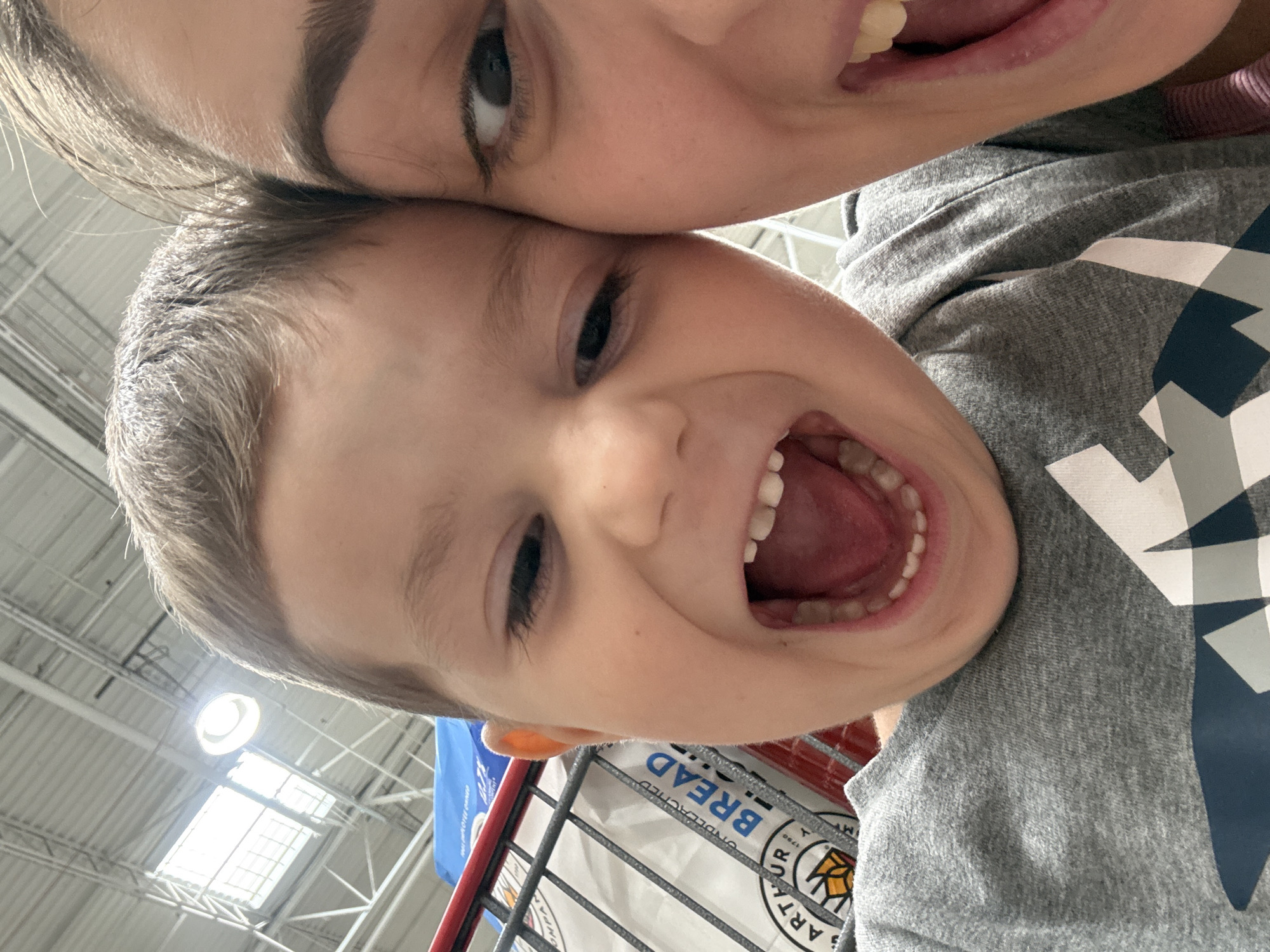 In an indoor setting, possibly a grocery store, a close-up selfie captures a young child and an adult. The child, positioned in the foreground on the left, has an open mouth, revealing teeth and the inside of their mouth, with their eyes wide. To the right, the adult's face is partially visible, with one eye open and looking towards the camera, and their mouth also open. Both individuals are wearing grey shirts. Behind them, a shopping cart is visible, containing a large bag of what appears to be flour, with "ARTHUR 1790" and "BREAD" printed on it. The background shows industrial-style ceiling structures and lighting, consistent with a large retail space. The location is the City of Yonkers, United States of America.