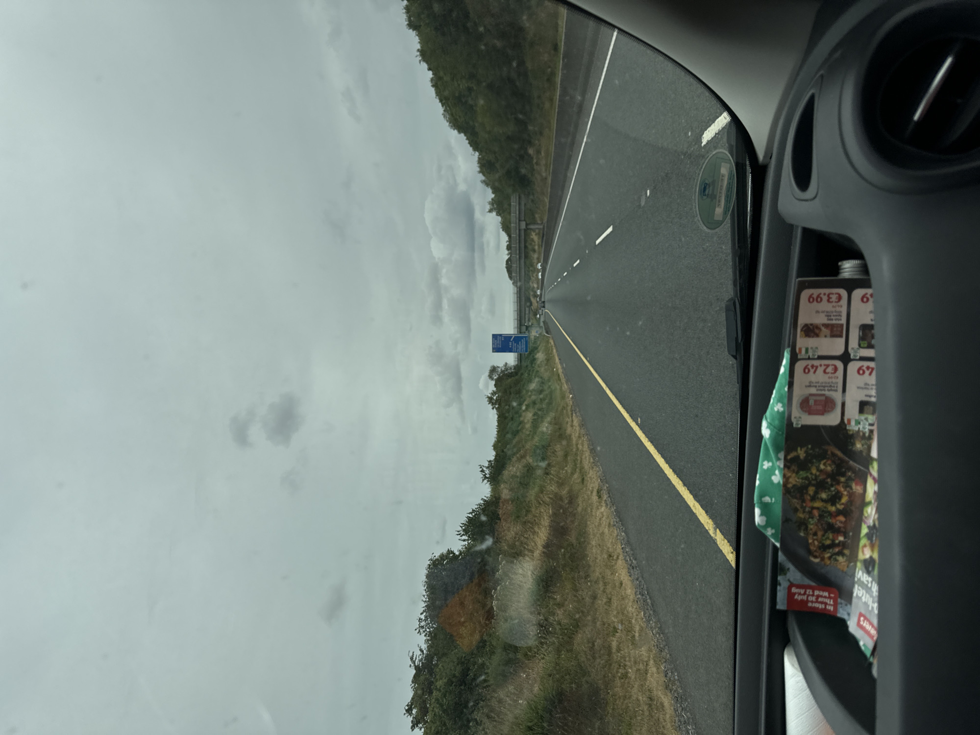 The image shows a view from inside a vehicle driving on a multi-lane highway. The highway is asphalt with a yellow line marking the left lane and white lines separating lanes. The sky is overcast with grey clouds. On the left side of the highway, there is a grassy embankment with bushes and trees. On the right side, there are also trees and a barrier. In the distance, a bridge crosses the highway, and there are other vehicles on the road.

Inside the vehicle, on the dashboard, there are several items including what appears to be a roll of paper towels on the left, and some leaflets or flyers are visible in a compartment to the right. One flyer has an image of food and prices in Euros, with text indicating "In store Thu 30 July - Wed 12 Aug" and prices such as "€2.49" and "€3.99". There is also a green fabric with a shamrock pattern visible. A circular sticker is affixed to the inside of the windshield.

The scene is consistent with being on a highway in Ireland, as indicated by the context of Linkardstown Lane. The overall impression is one of a typical highway journey on a cloudy day.