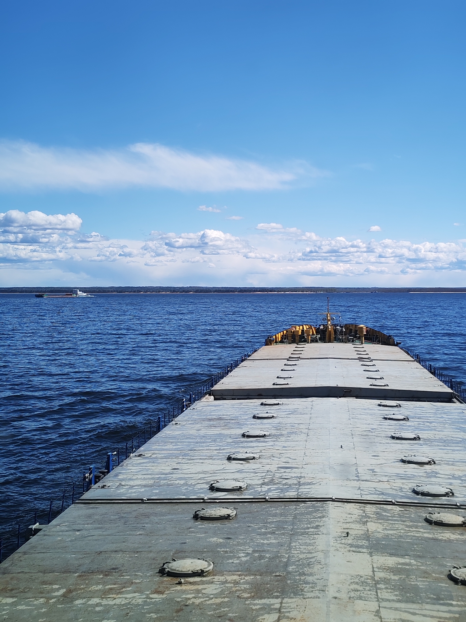 A low-angle, eye-level photograph depicts the deck of a barge, likely on the Volga River near Ozernoe Ust'e, Russia. The barge deck, with numerous circular hatches, stretches towards the center horizon. Beyond the barge, a wide expanse of dark blue water extends to a distant shoreline, lined with trees. On the left horizon, a small tugboat pulls a barge. The sky above is a bright blue with scattered white clouds.
