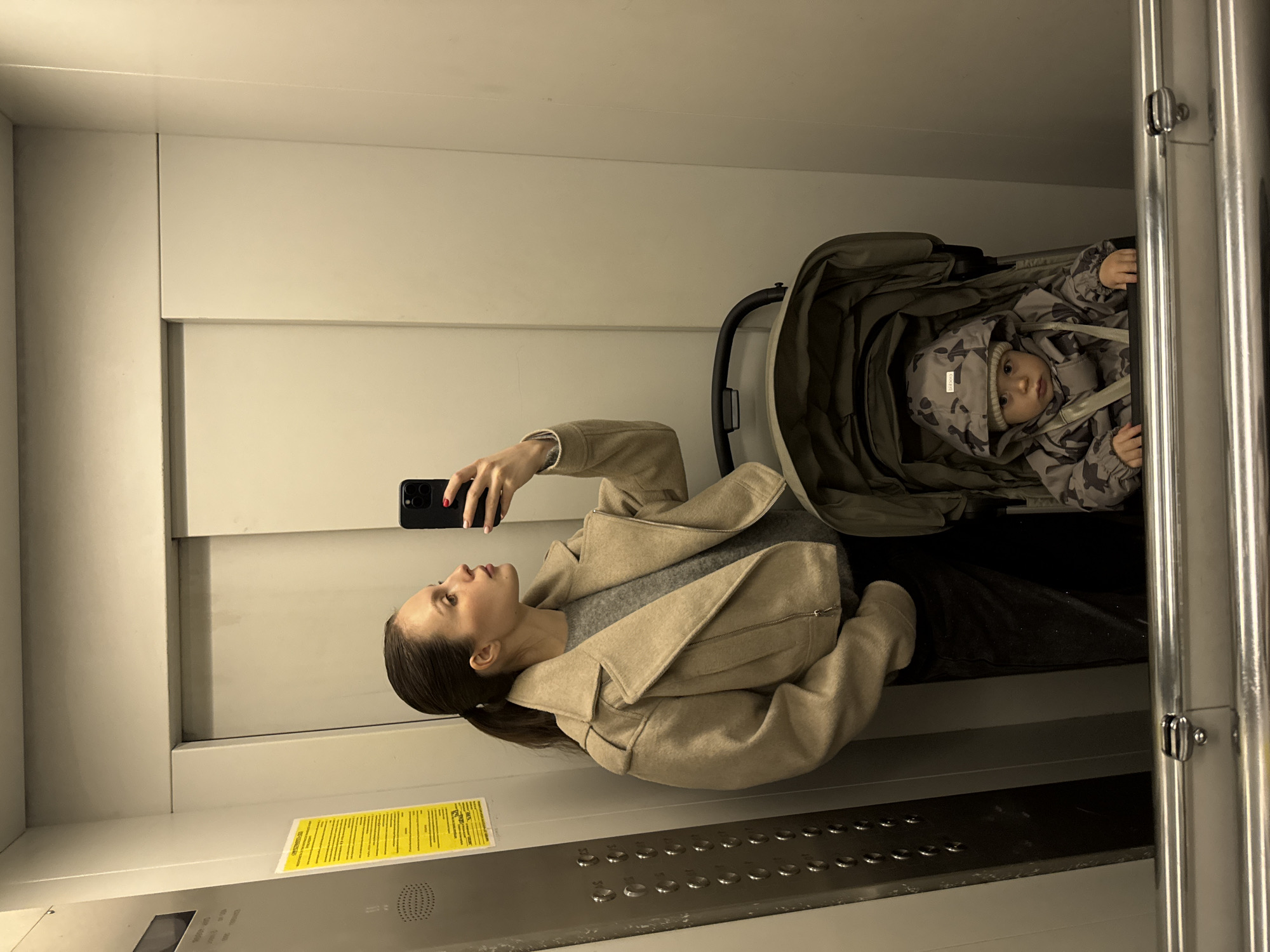 The image shows a woman with brown hair in a beige coat taking a selfie with her smartphone in an elevator. A baby in a stroller is next to her. The elevator has white walls and a control panel with buttons. A yellow information sign is visible. The location is Moscow, Russia.