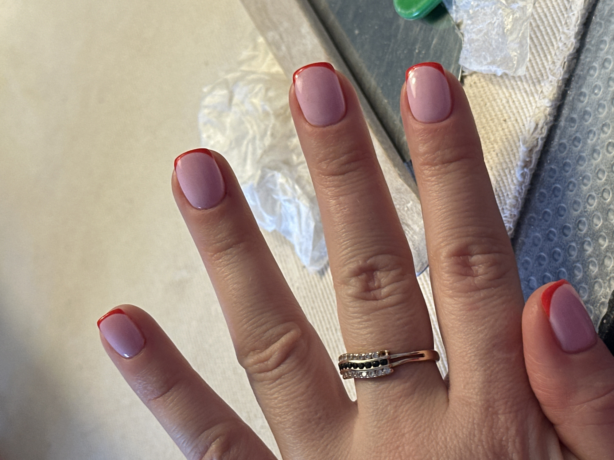 The image shows a close-up view of a woman's hand with neatly manicured fingernails. The nails have a pale pink base color with a vibrant red French tip. The woman is wearing a ring on her left ring finger, which appears to be a band adorned with small black and clear stones. The background consists of a textured surface, possibly a table or mat, with some white material, possibly cotton pads or tissue, and a gray object that could be a nail file. The lighting suggests an indoor setting. There is no discernible information about the location within Nevinnomyssk, Russia.