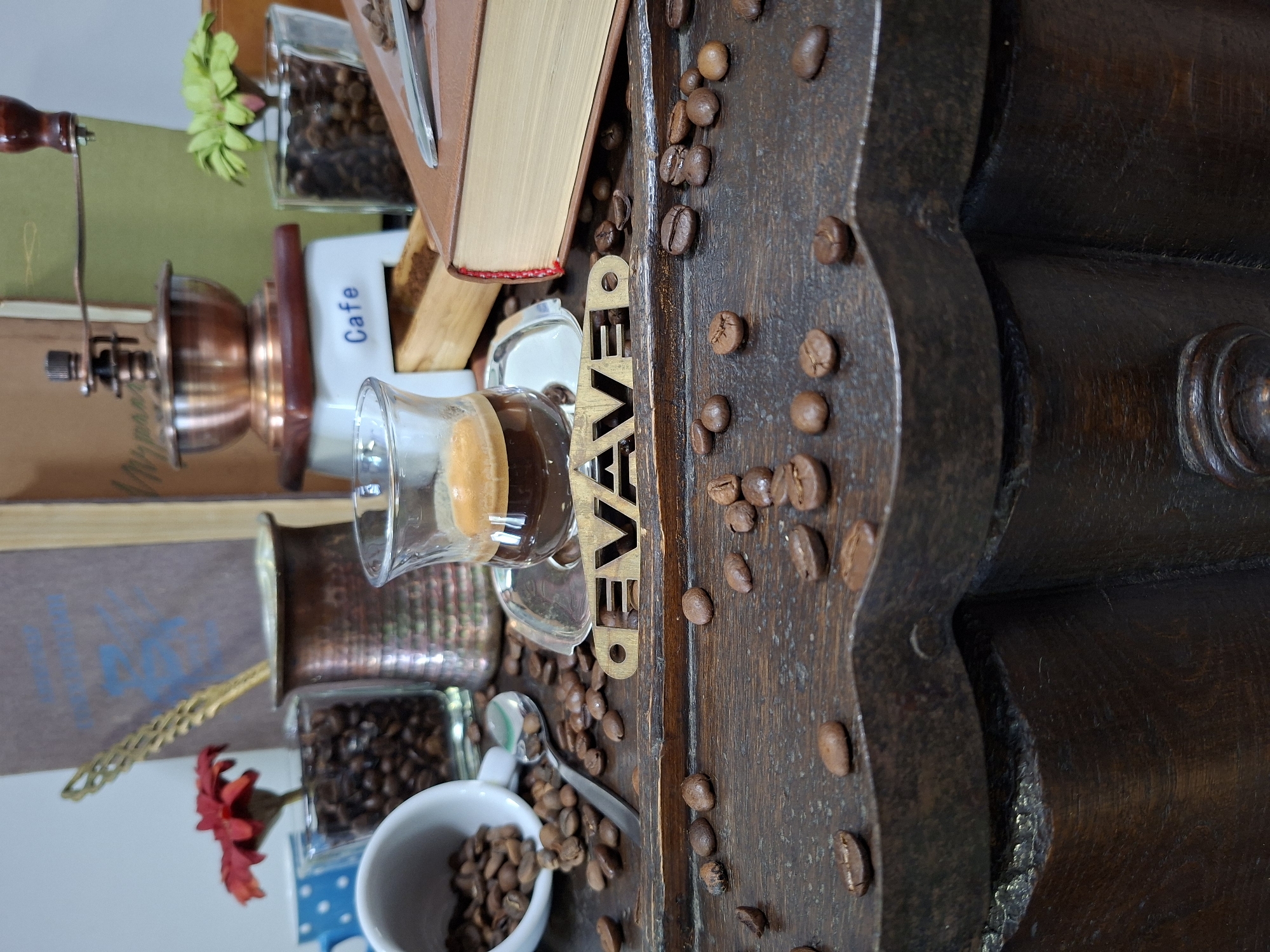 A still life photograph is captured at a low angle, focusing on a dark wooden surface adorned with scattered coffee beans. In the foreground, a decorative wooden cutout spelling "CAVE" is partially visible. The midground features a glass of espresso with a rich crema, resting on a silver saucer. To the left, a white teacup is overturned, spilling coffee beans onto the wooden surface. Behind the espresso, a small, decorative copper pot sits near a glass container filled with coffee beans and a red artificial flower. Further back, a vintage copper coffee grinder with a wooden base is positioned in front of a stack of books. To the right, another glass container holds coffee beans and a small green plant. A closed brown book lies on the right side of the frame. The background consists of blurred books and a light-colored wall. There are no visible people or actions. The scene evokes a coffee-themed setting, possibly in a cafe or home environment. There is no information available to determine the location in Chișinău, Moldova.