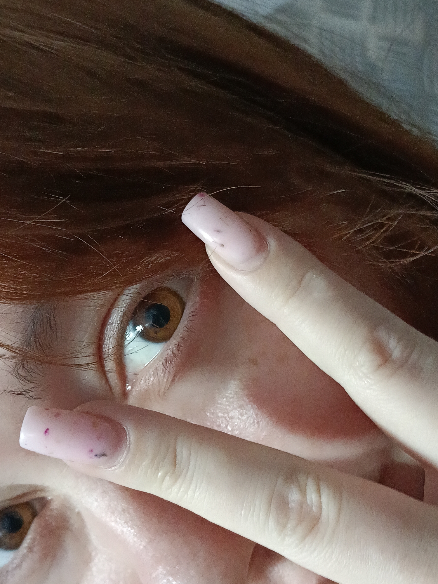 A close-up, slightly angled view of a person's face, primarily focusing on one eye. The eye is brown and partially obscured by two fingers, which are light-skinned with squared, pale pink manicured nails. The fingers form a "peace sign" gesture, positioned in front of the face. The person has reddish-brown hair visible in the upper and left portions of the frame. The background is indistinct and appears to be a neutral color. No specific location within Chaikovsky, Russia, can be determined from the image. No specific actions or interactions other than the posed gesture are evident.