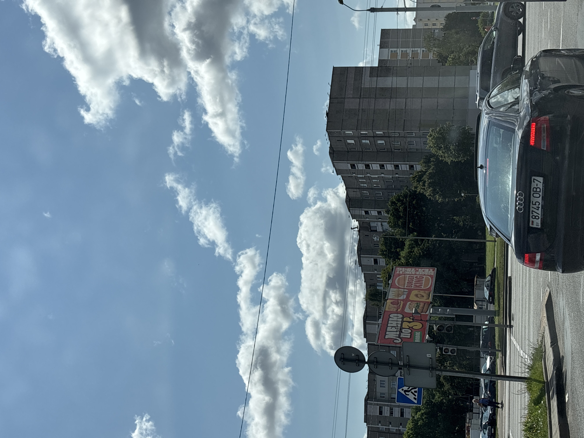 In Minsk, Belarus, on a bright, partly cloudy day, a street view captures a bustling urban intersection. The foreground features a black Audi sedan, its license plate reading "BY 8745 OB-7". To its left, a dark gray Audi SUV is visible. Further into the scene, a pedestrian, a woman with blonde hair wearing a light blue top and dark pants, walks across a crosswalk. A billboard advertising Burger King is prominently displayed near the intersection, showing menu items. Traffic lights and street signs are present, indicating a regulated intersection. In the background, several multi-story apartment buildings dominate the skyline, typical of Soviet-era architecture. Lush green trees line the streets, adding a natural element to the urban landscape. The sky is a clear blue, with scattered white clouds. The overall atmosphere suggests a typical day in a Belarusian city.